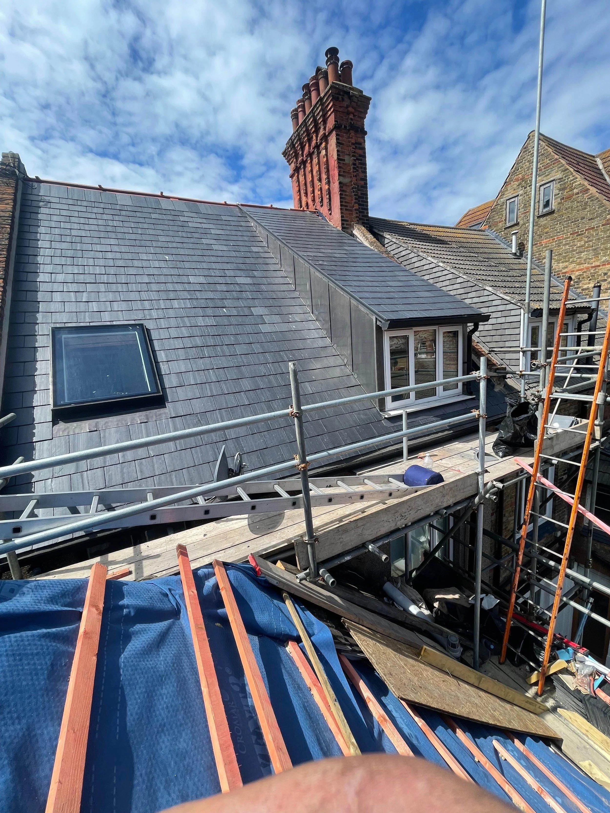 View of a house roof under construction, with scaffolding, a skylight window, and a chimney against a cloudy sky.