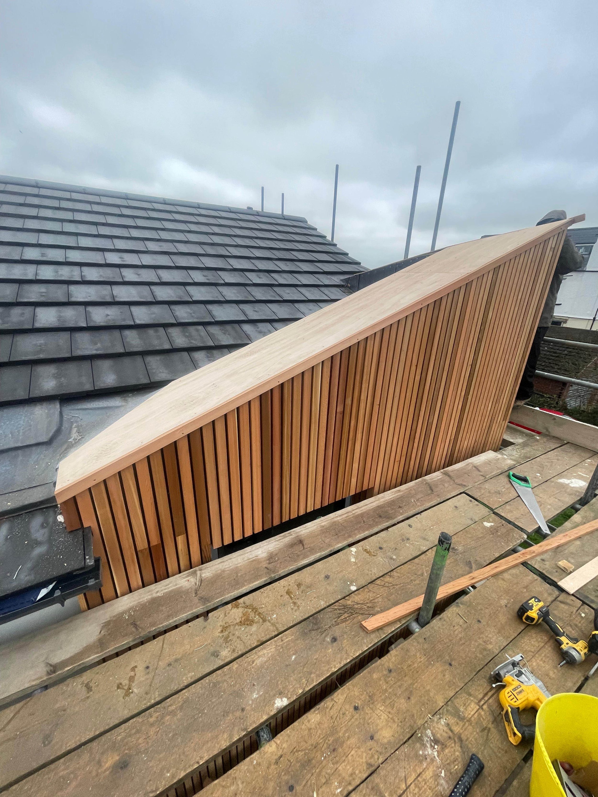 A construction site showing a worker installing vertical wooden slats on a sloped roof section with gray shingles, using tools like a saw and drill, on a cloudy day.