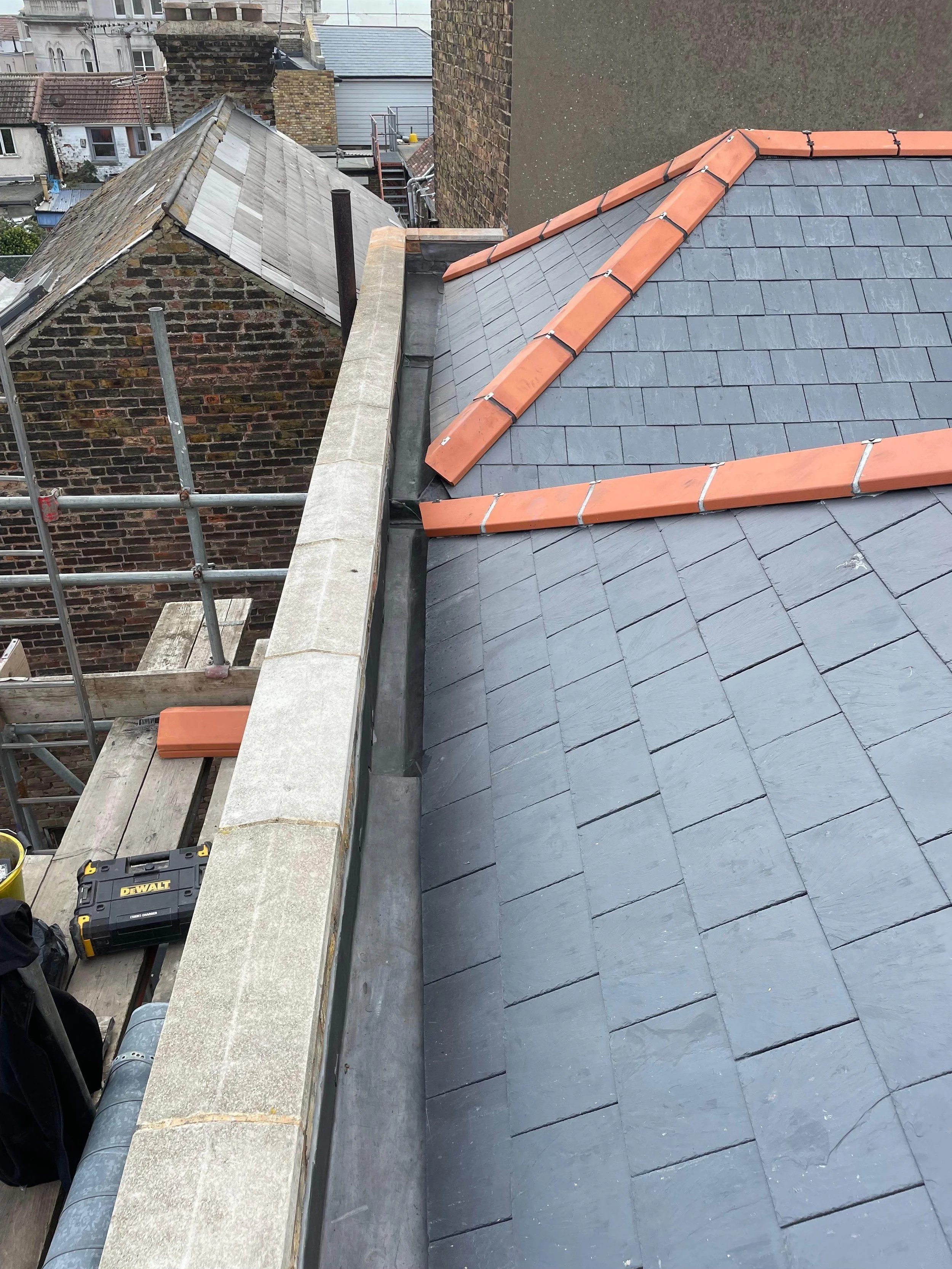 View of residential rooftops under construction, showing slate roofing, brick chimneys, scaffolding, and construction tools.