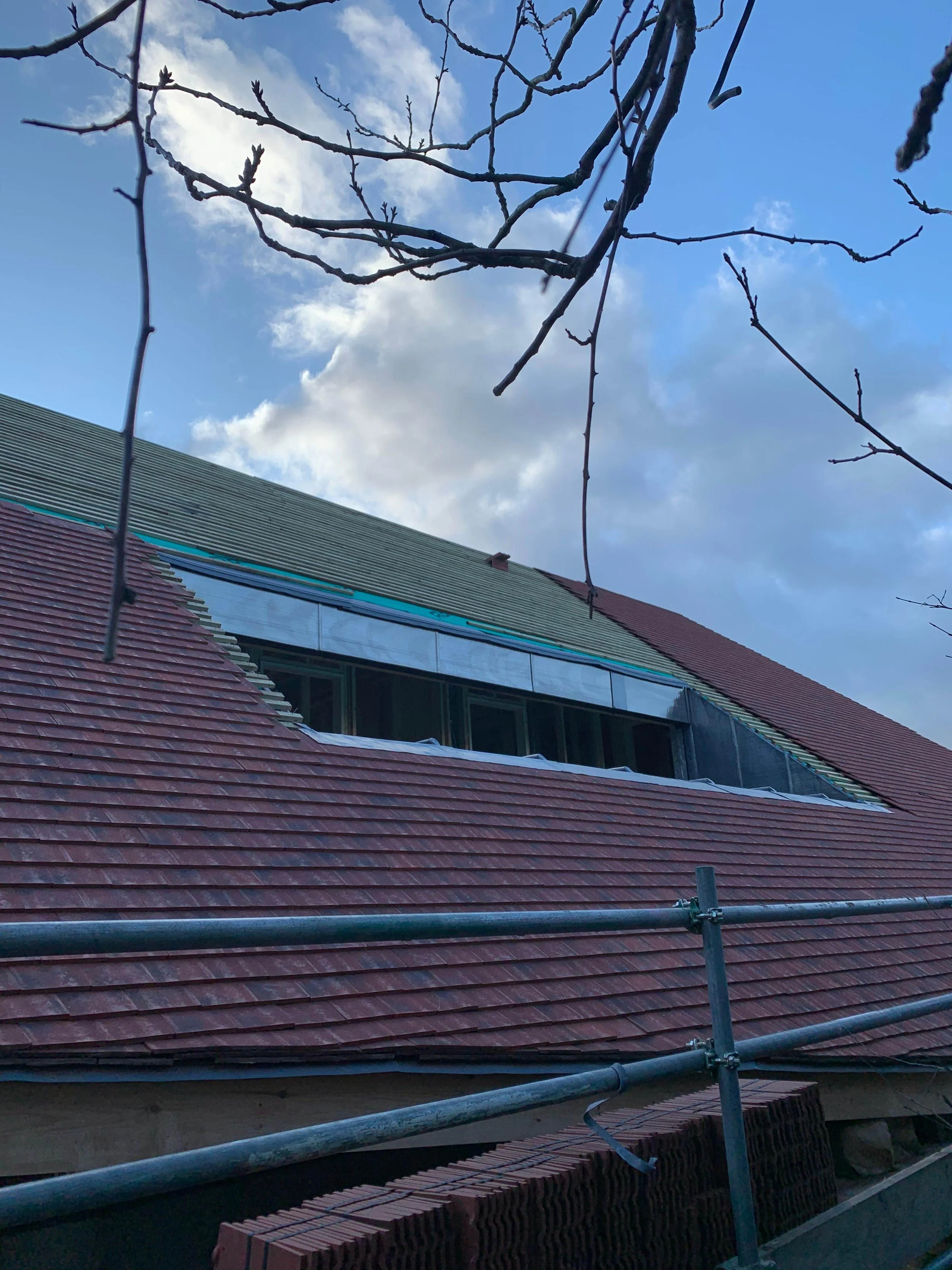 Part of a building under construction or renovation, with a sloped roof covered with red tiles, scaffolding in the foreground, and bare tree branches at the top, with a partly cloudy sky in the background.