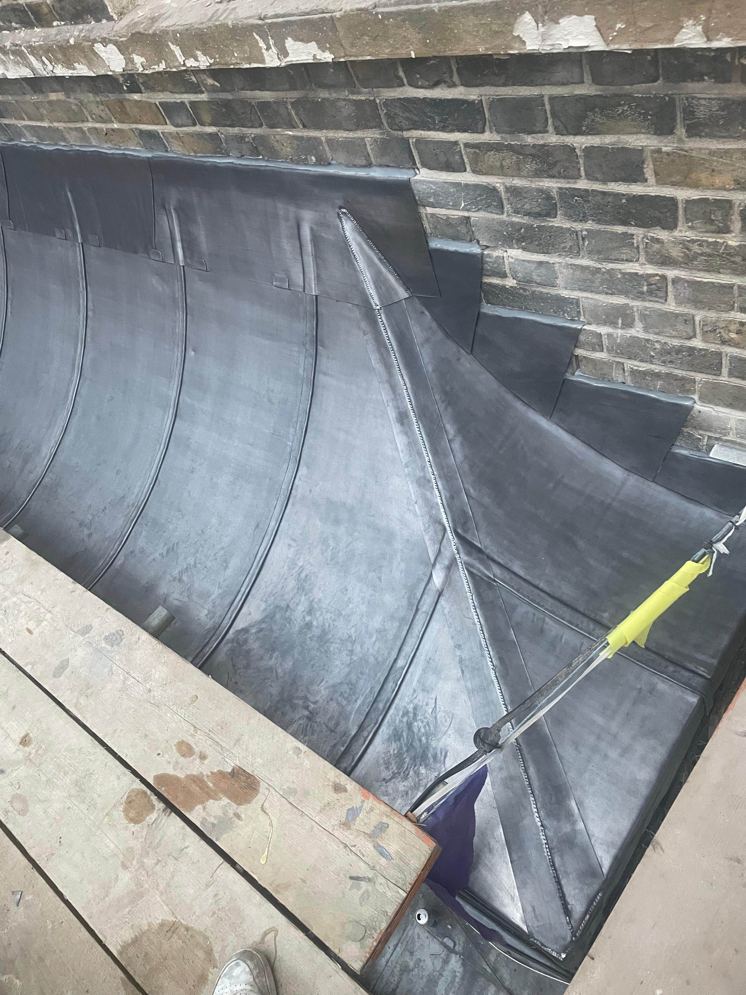 Close-up of a metal roofing sheet with a zipper-style seam, installed against a brick wall with some wooden planks and part of a person's shoe visible at the bottom.