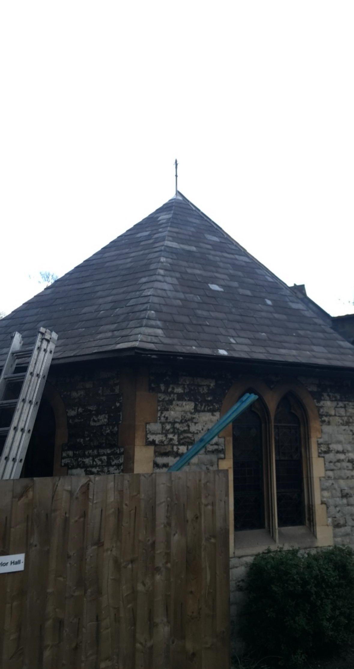 A small stone church with a steep roof, a weather vane at the top, and arched windows, partially obscured by a wooden fence and a bush, with a ladder resting against the side of the church.
