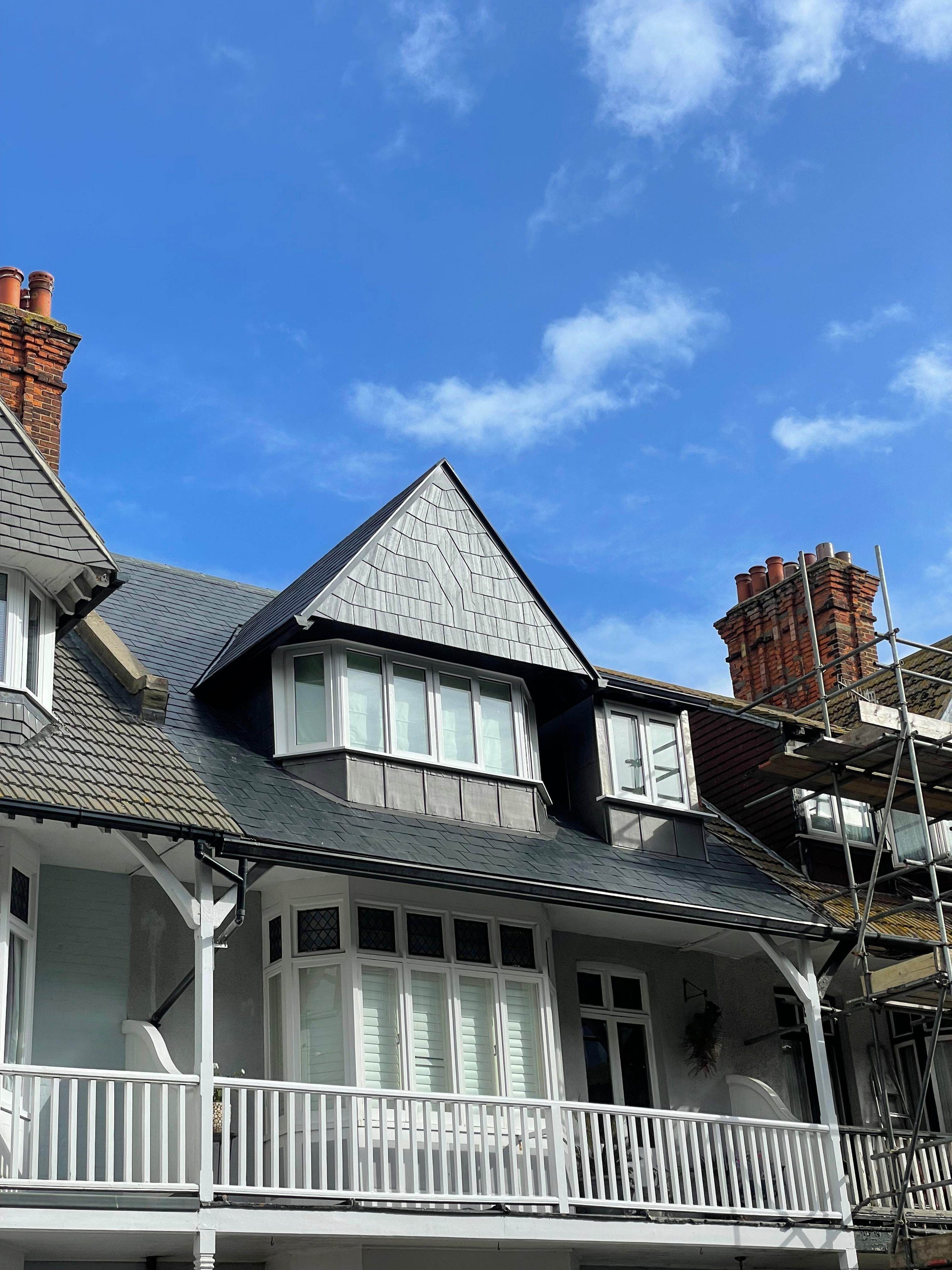Close-up of a multi-story house with a steep roof, dormer windows, a white balcony, and scaffolding on one side, against a bright blue sky with scattered clouds.