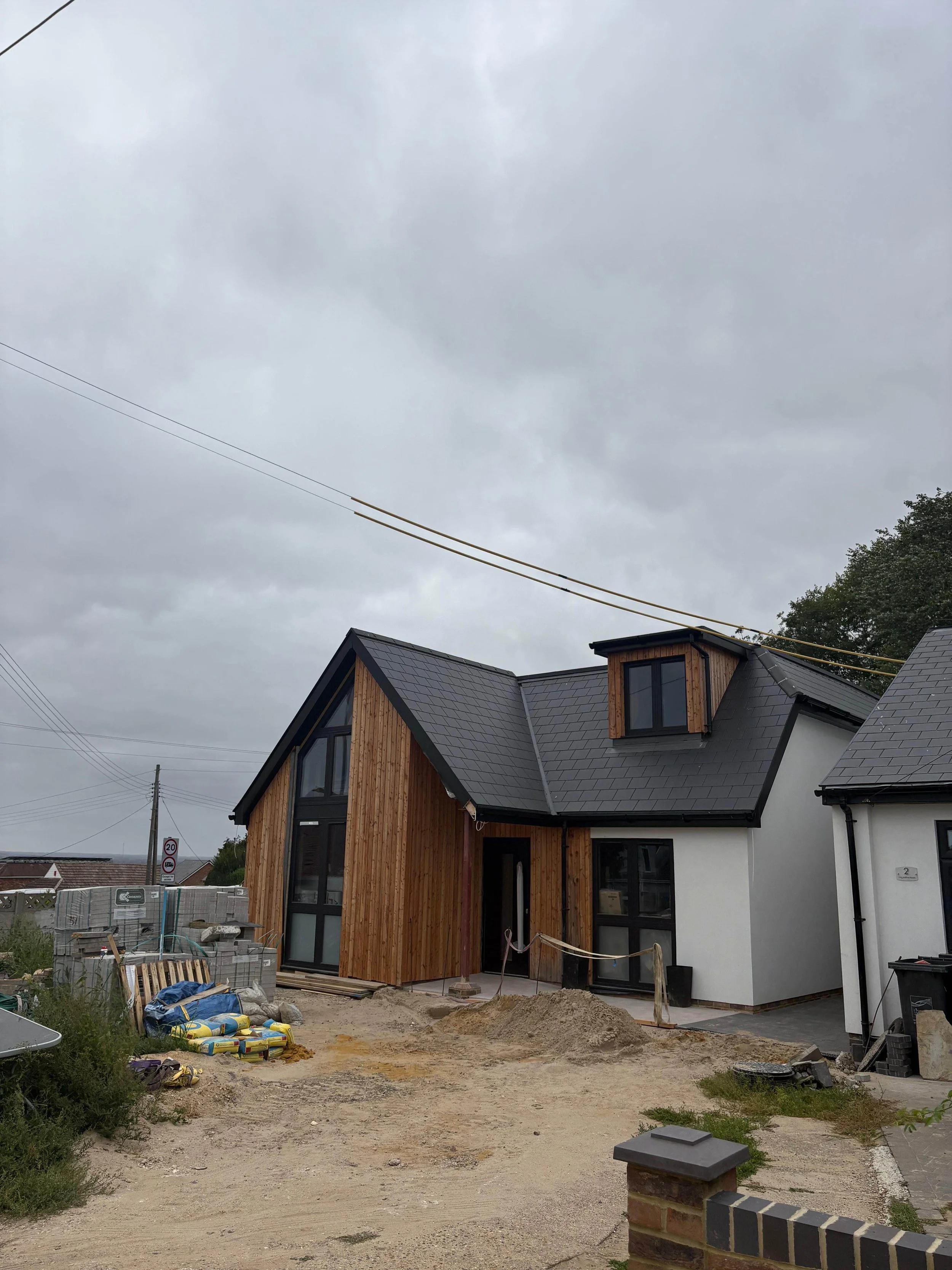 Under construction house with black roof and wooden and white exterior, construction materials and dirt in front, overcast sky.