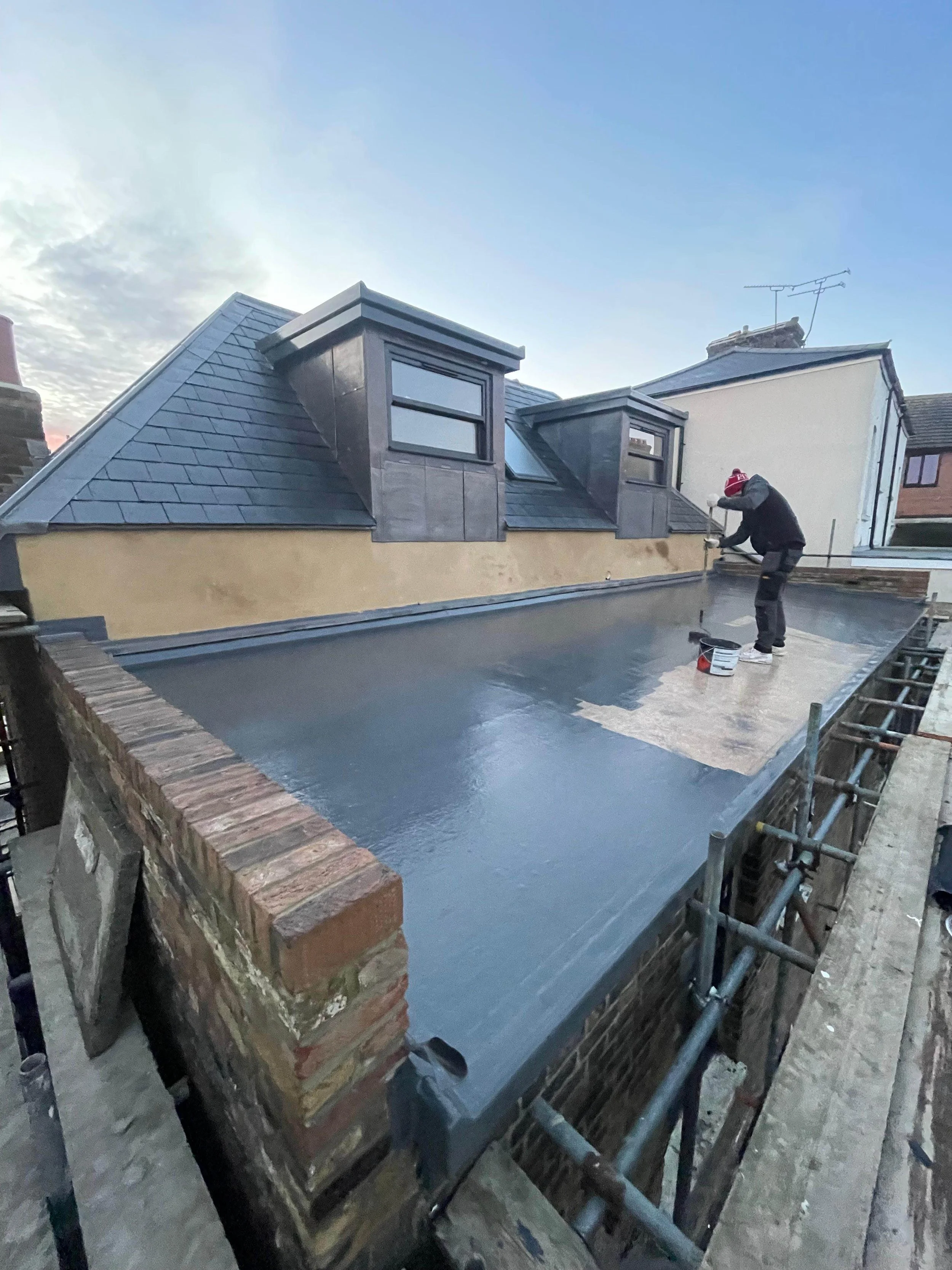 A worker applying a waterproof coating to a flat rooftop of a building, with neighboring buildings visible under a clear sky.
