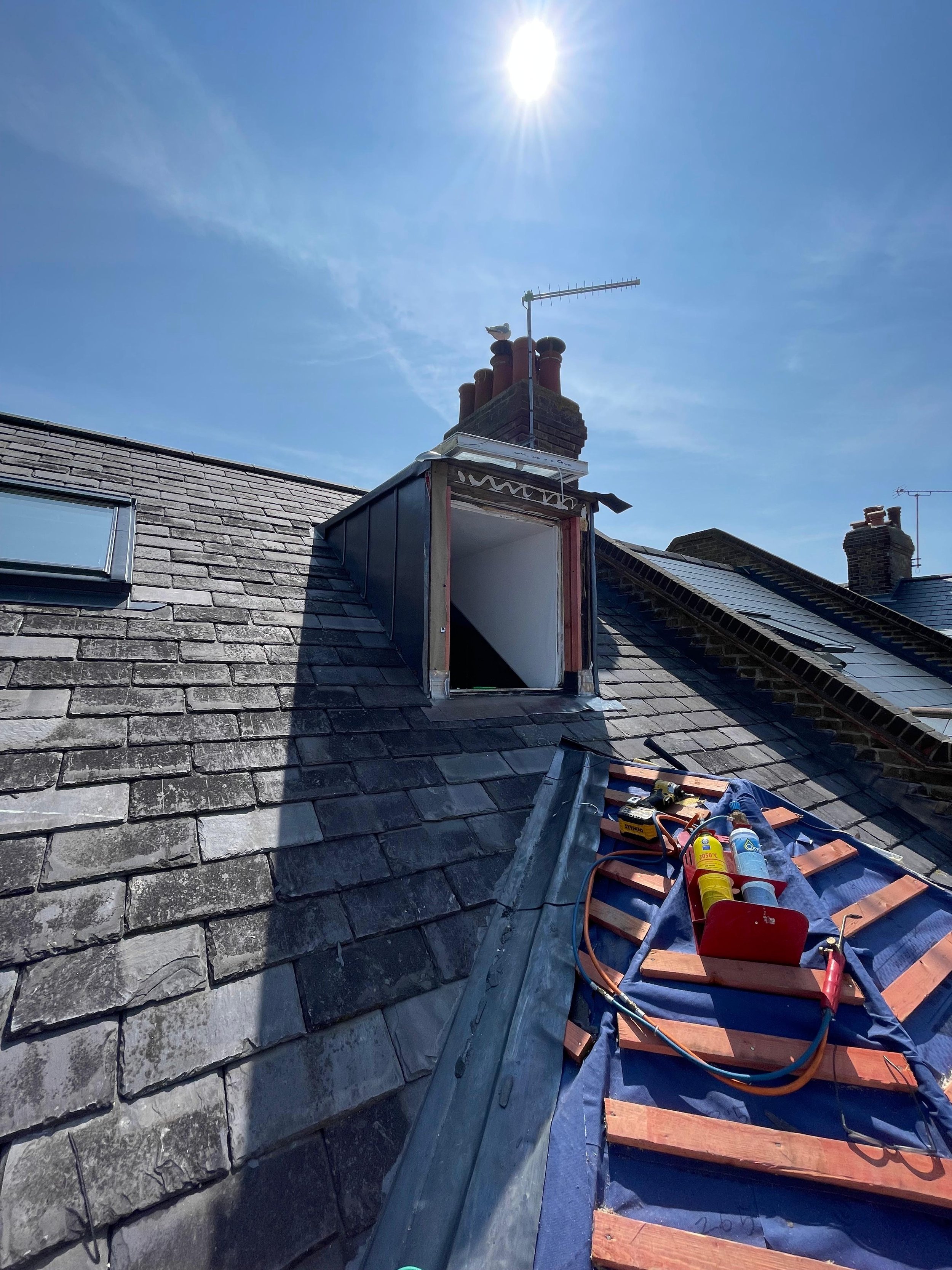 Roof of a building under repair with tools, wooden battens, and a blue tarp, and a chimney with an antenna against a bright blue sky.