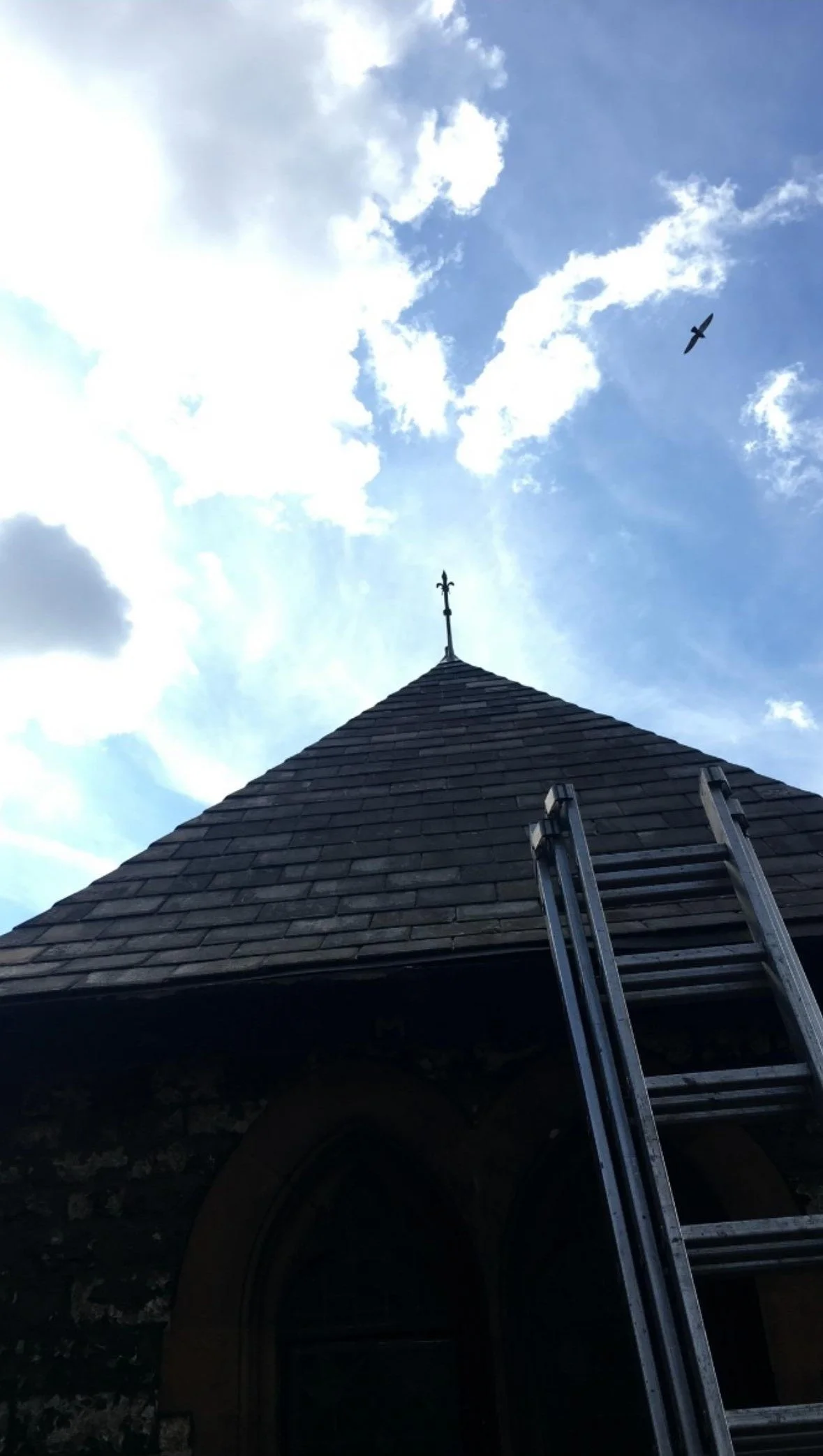 View of a church steeple with a ladder leaning against it, under a partly cloudy sky with two birds flying.