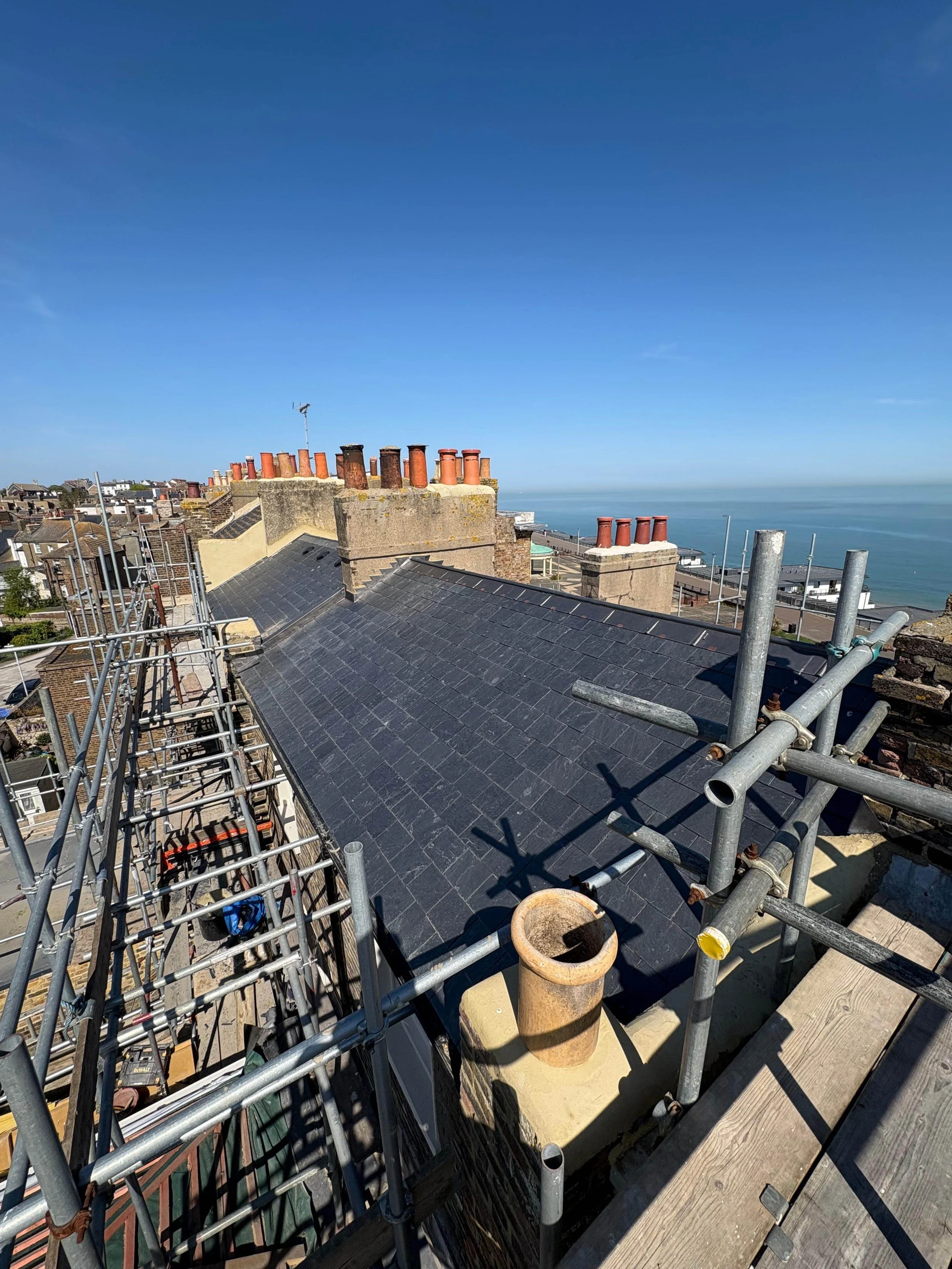 View of rooftops with scaffolding, chimneys, and the ocean in the distance under a blue sky.