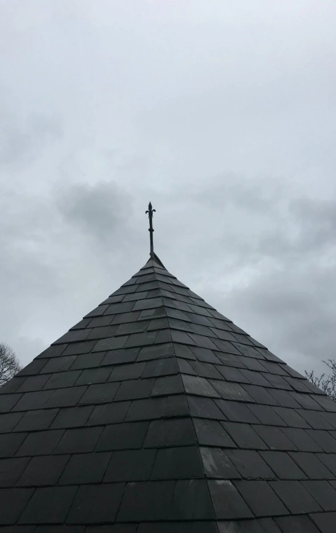 View of a slate tiled roof with a metal cross on top, against a cloudy gray sky.
