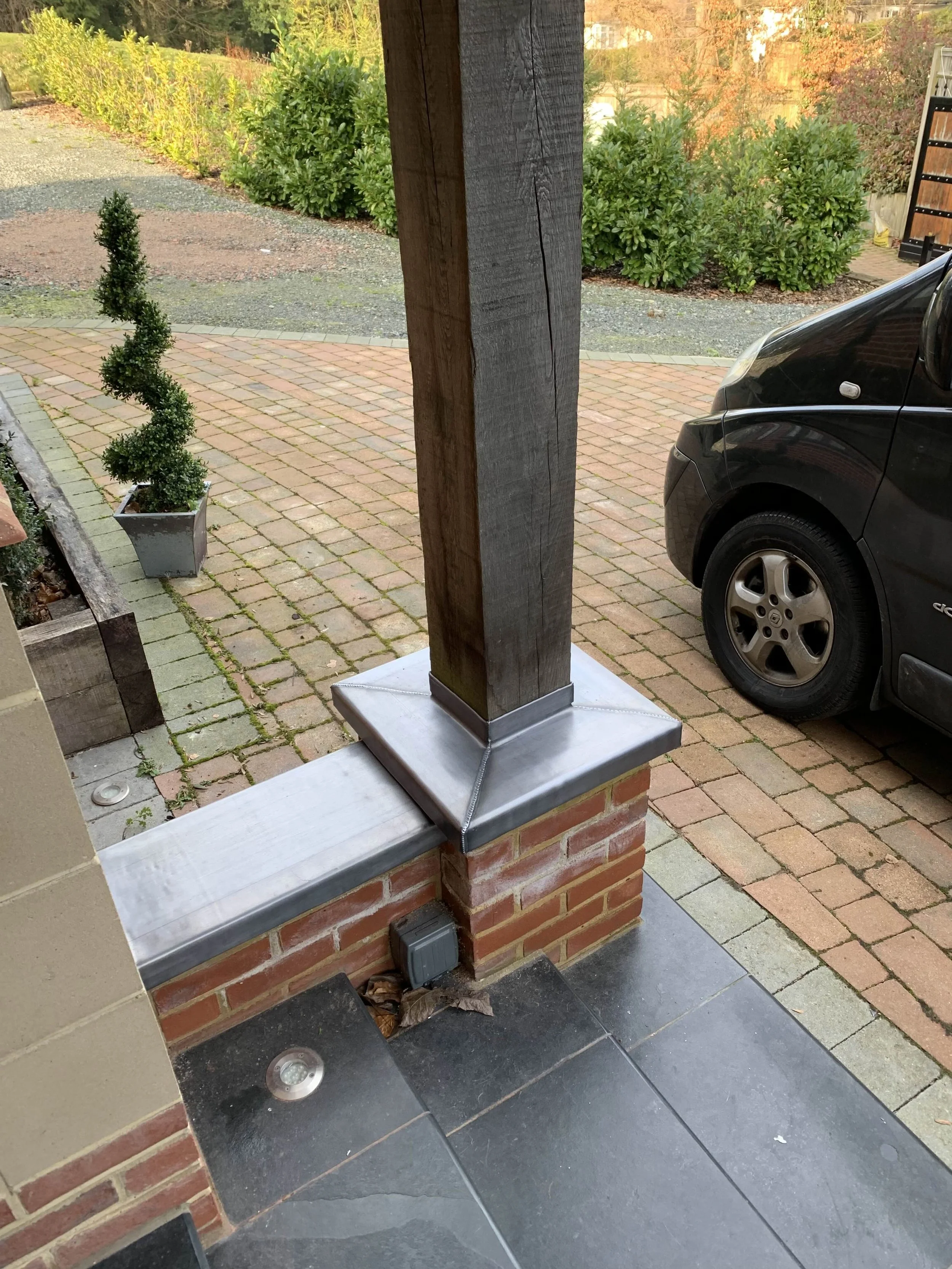 Close-up of a brick porch with a wooden post and metal post base; a black car is parked nearby on a brick driveway, with shrubs and trees in the background.