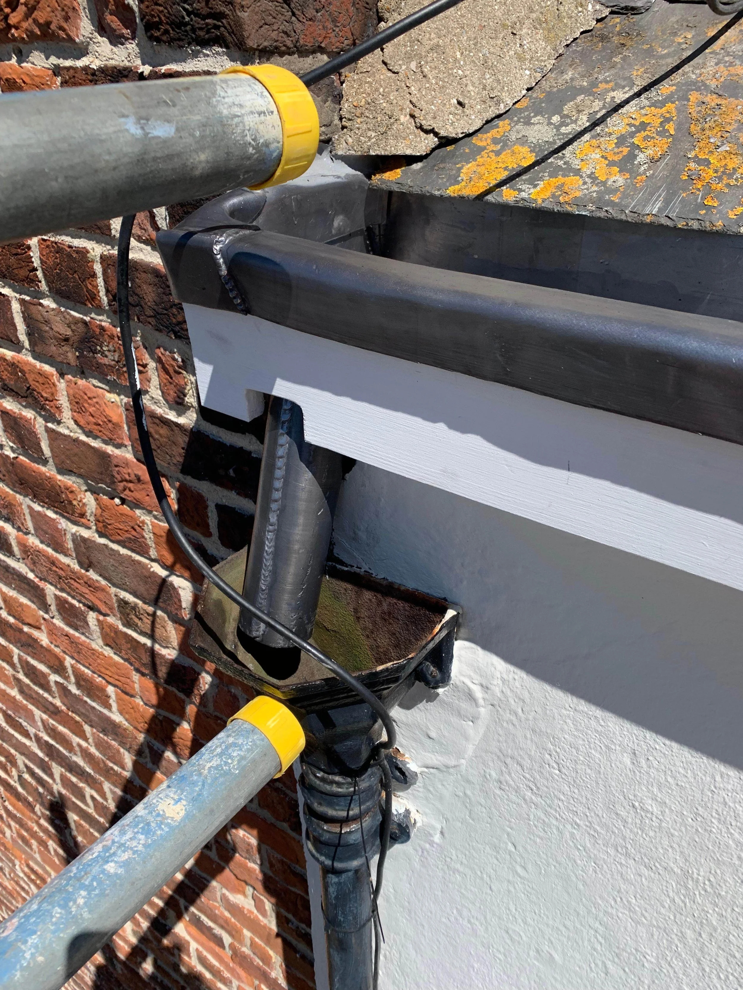 Close-up view of building construction with metal flashing, white wood trim, brick wall, and part of a gutter system, with metal pipes and wires attached.