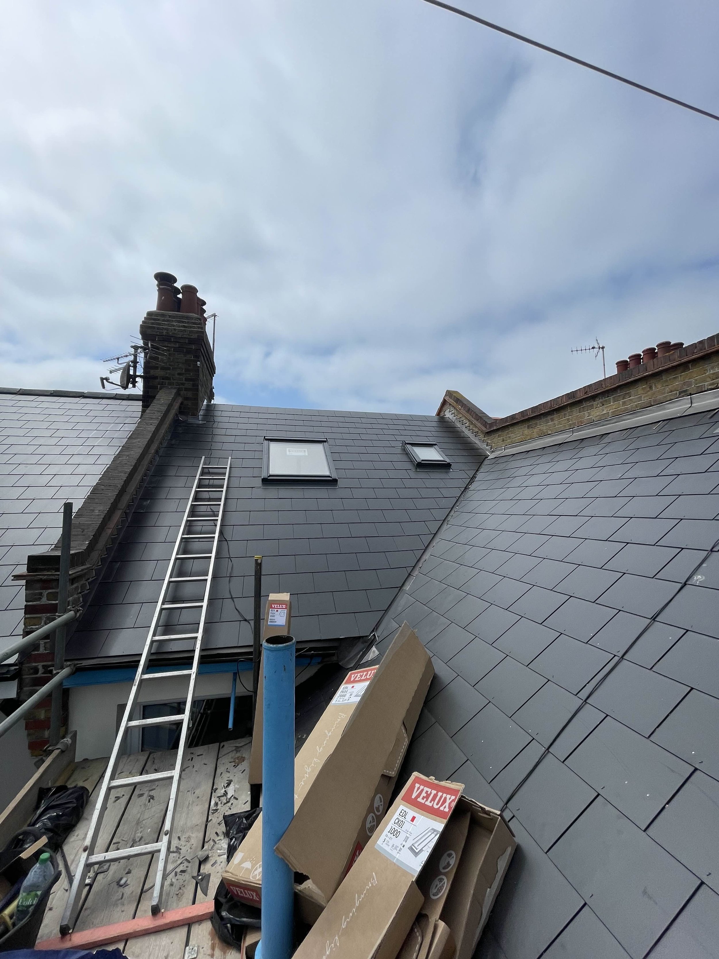 View of a house roof with newly installed dark grey slate tiles, windows, ladder, and construction materials in the foreground, under a cloudy sky.
