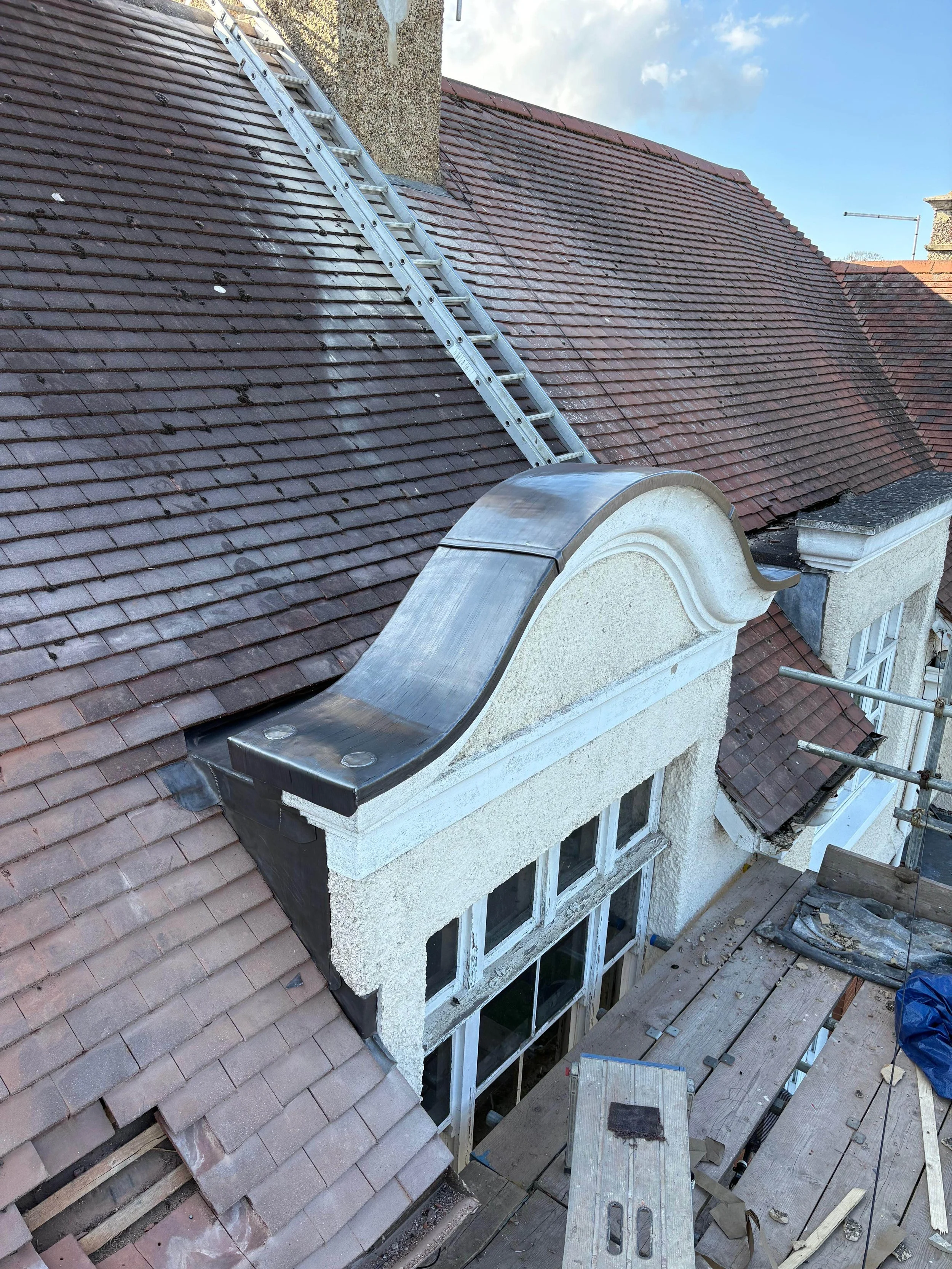 Construction work on a house roof with scaffolding, showing a decorative curved metal feature on the front of the roof and scaffolding platform on the side.