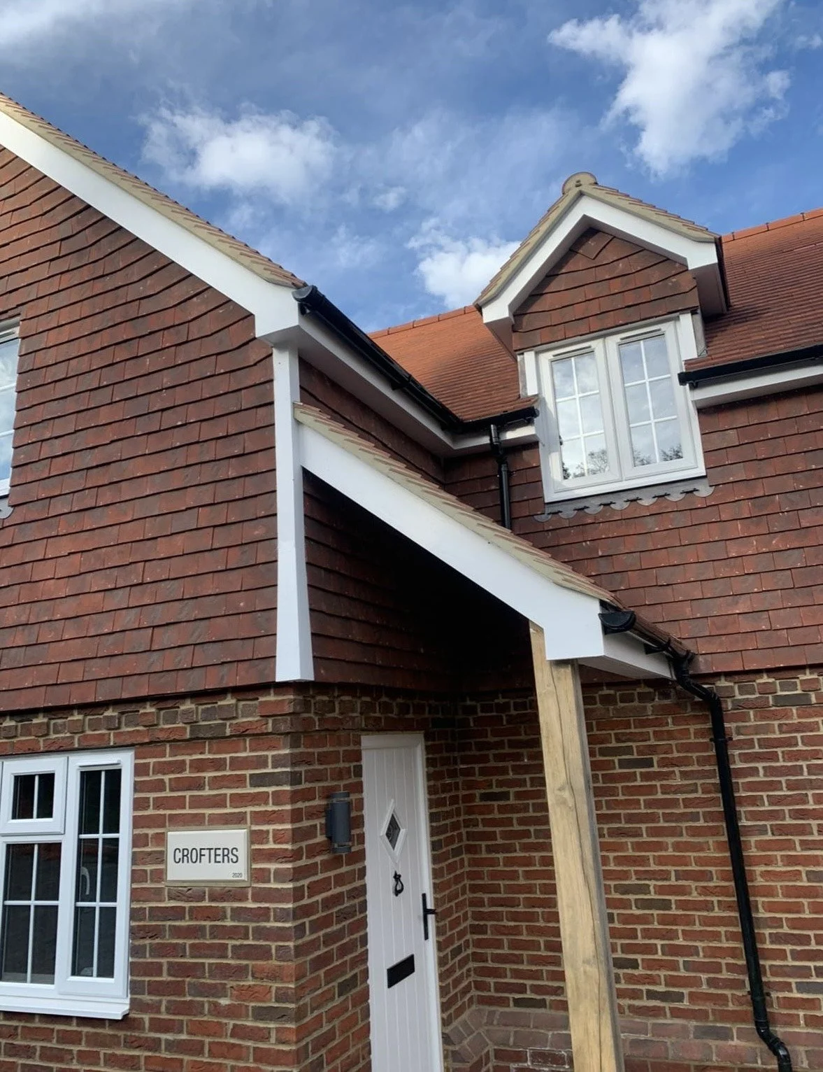 Close-up of a brick house with white window frames, black rain gutter, sign that reads 'CROFTERS,' and a new wooden support post near the entrance, under a blue sky with clouds.