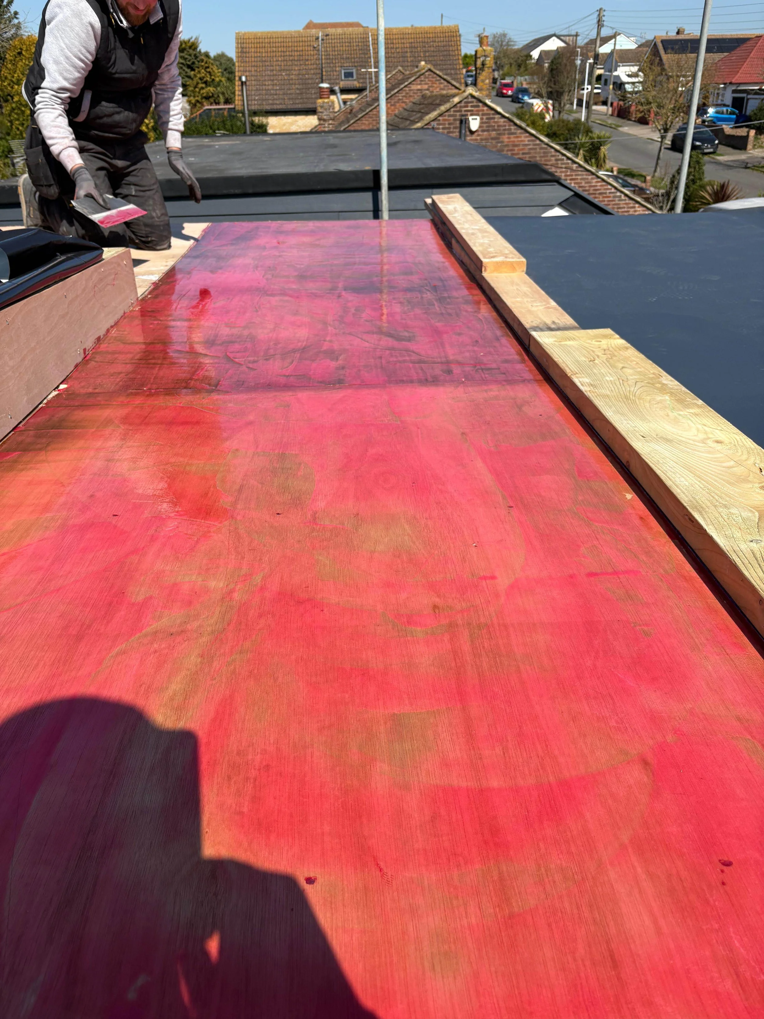 A person working on a flat rooftop, painting or staining a large wooden panel red, with houses and a street visible in the background.