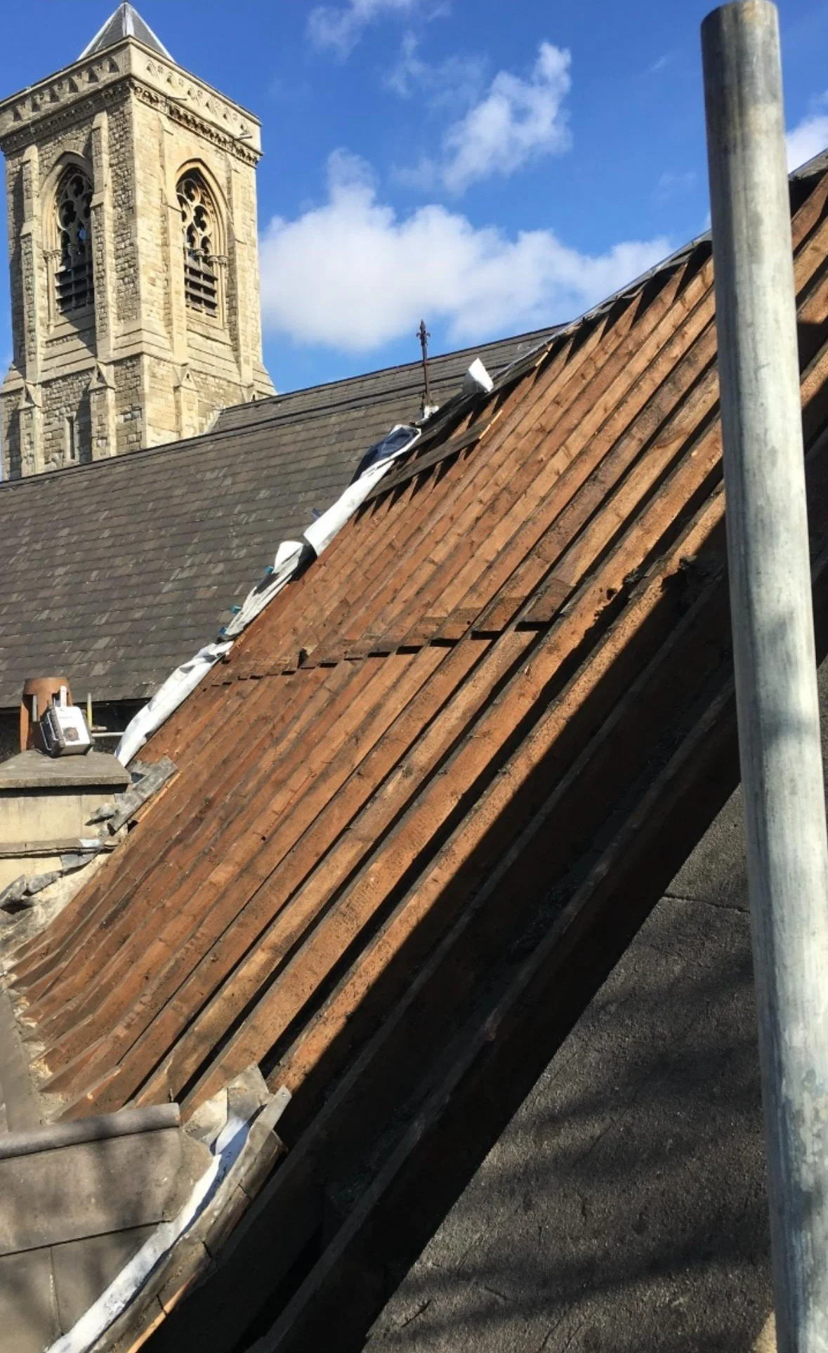 Side view of a roof under renovation, with some tiles removed and scaffolding visible, with a church steeple in the background and a partly cloudy sky.