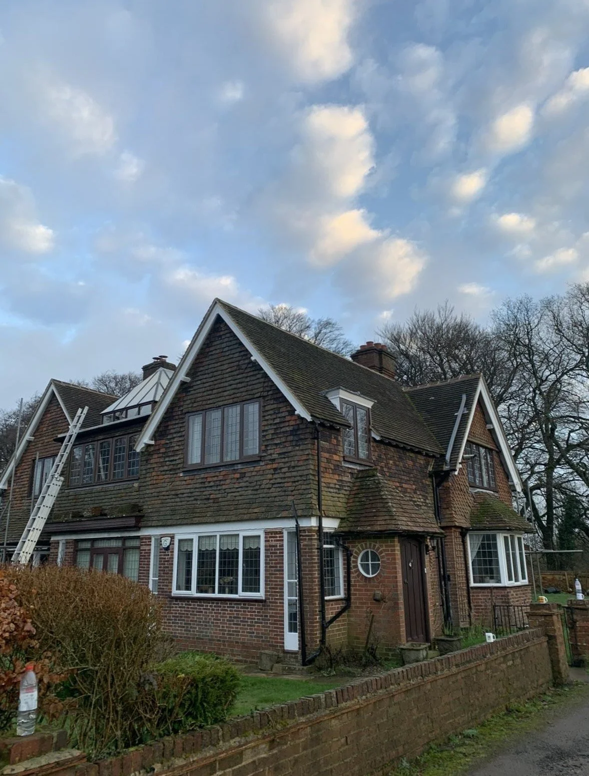 A large brick house with a steep gabled roof, multiple windows, and a small circular window near the entrance. There is a ladder leaning against the house on the left side, and a brick wall borders the front yard. The sky is partly cloudy with patche