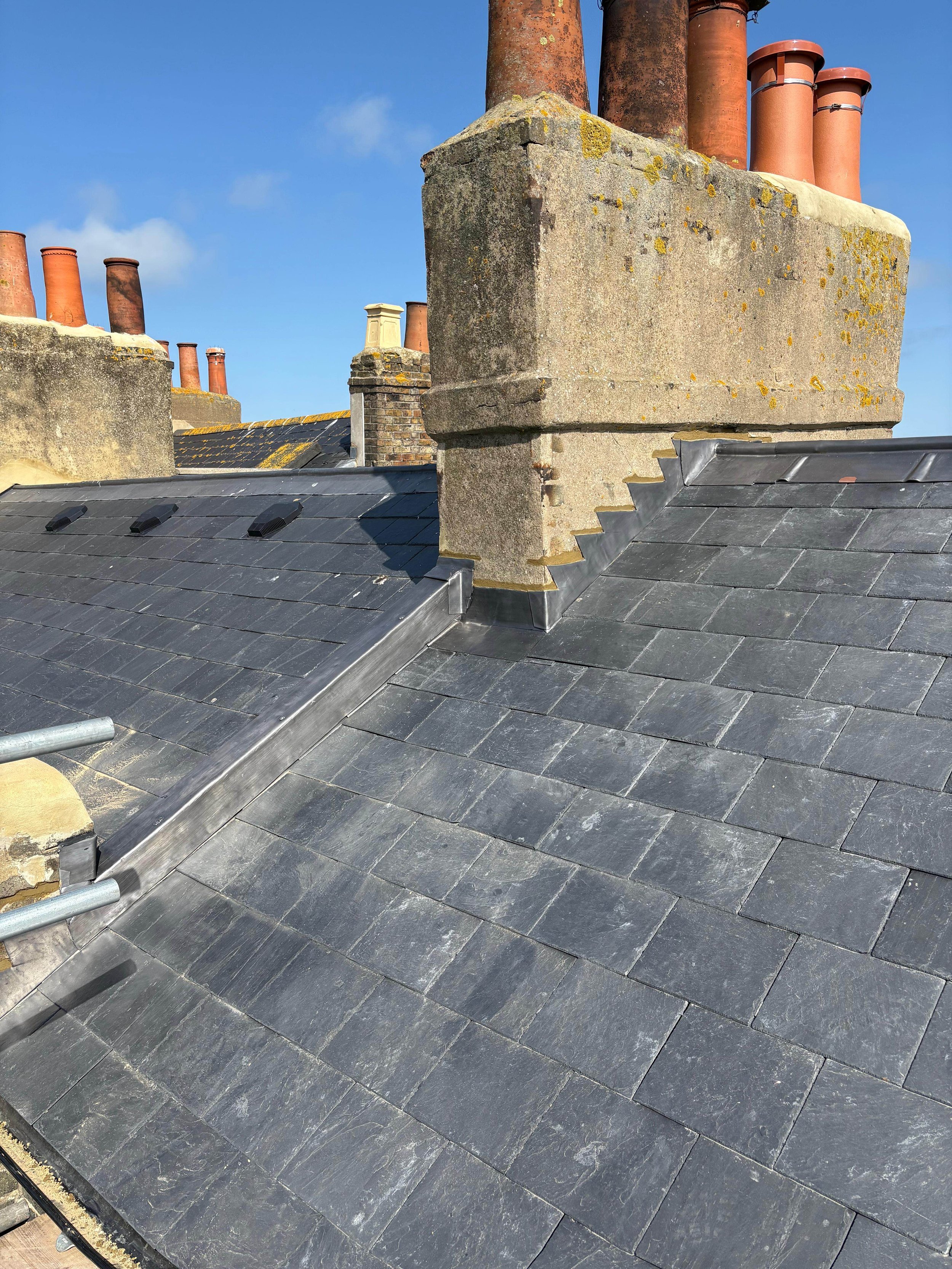 Close-up of a rooftop with slate tiles and brick chimney stacks against a blue sky.