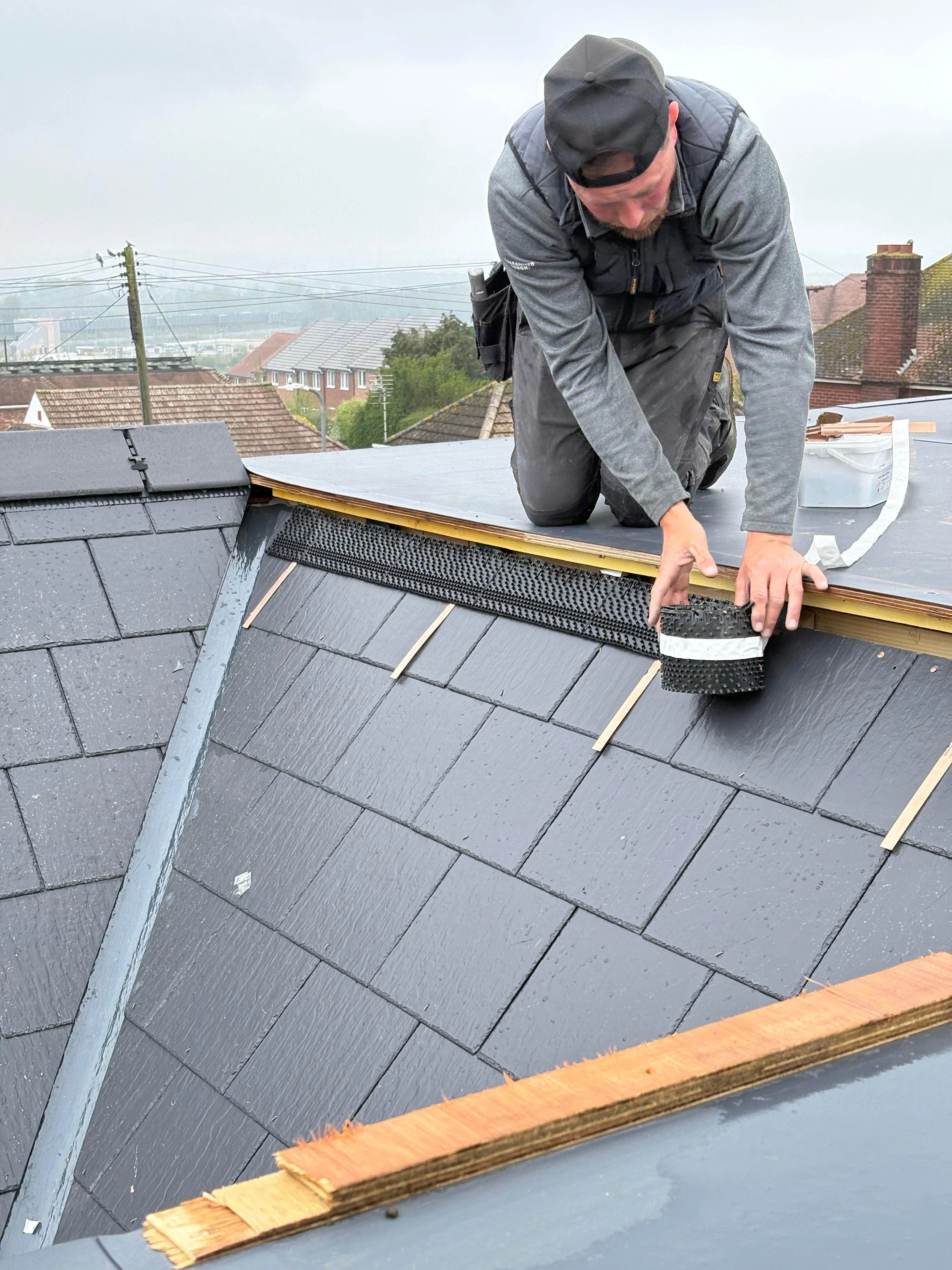 A man kneeling on a roof working on roofing materials, with rooftops and overcast sky in the background.