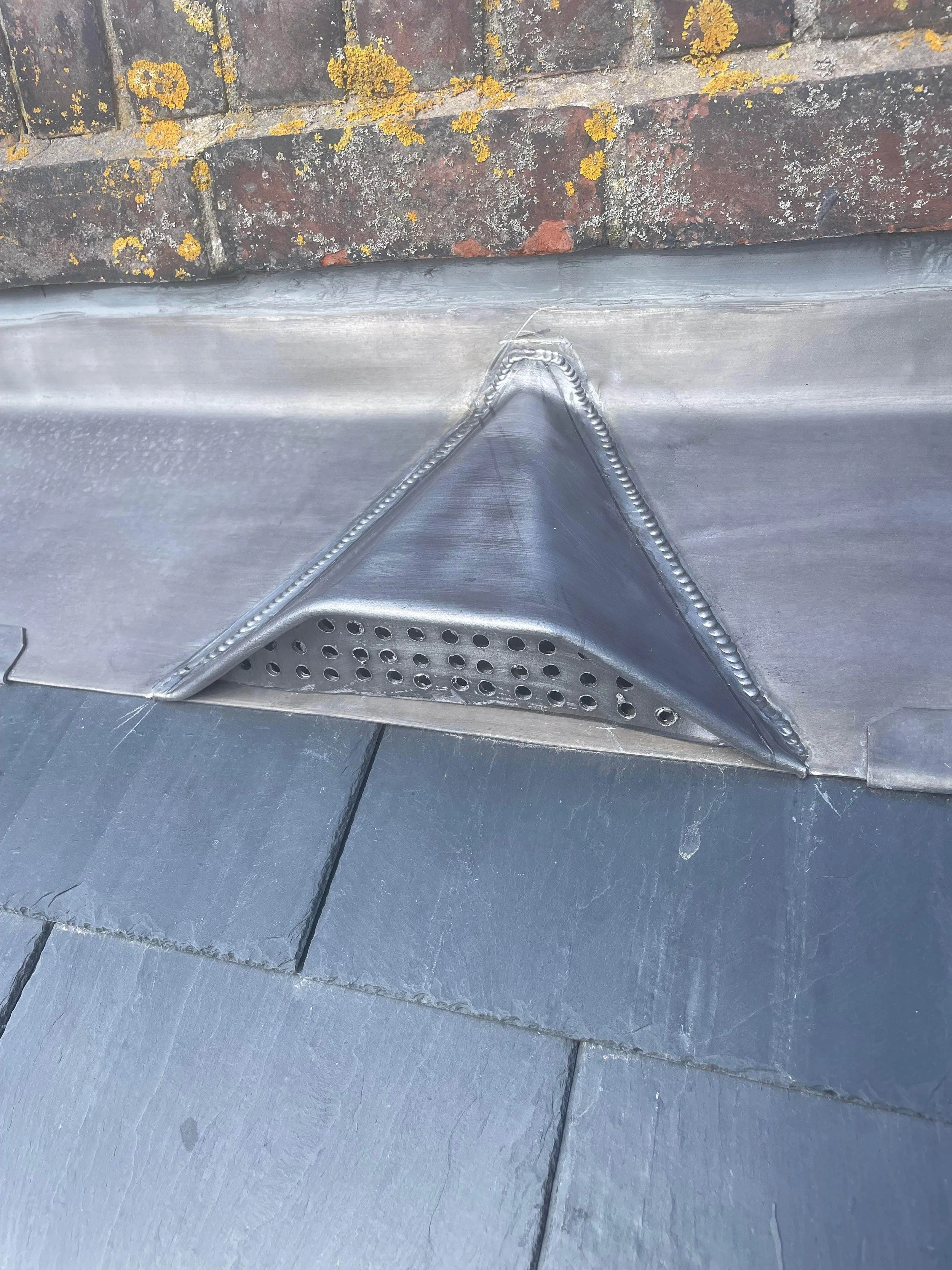 Close-up view of a metal roof vent with rounded base, mounted on slate tiles, with a brick wall in the background covered in yellow lichen.