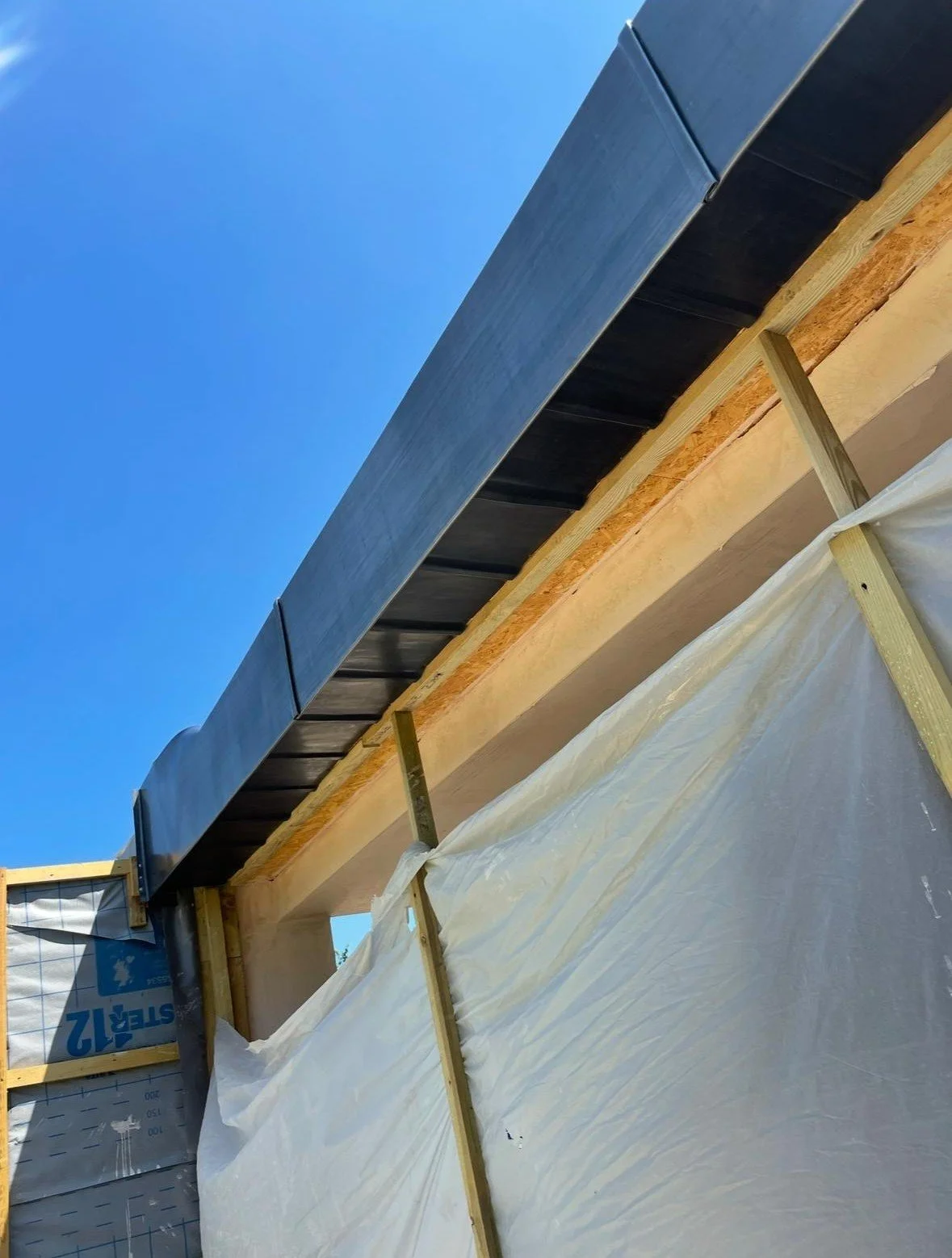 Construction site showing an overhang with black roofing material, supported by wooden beams covered with a white protective sheet, and a clear blue sky in the background.