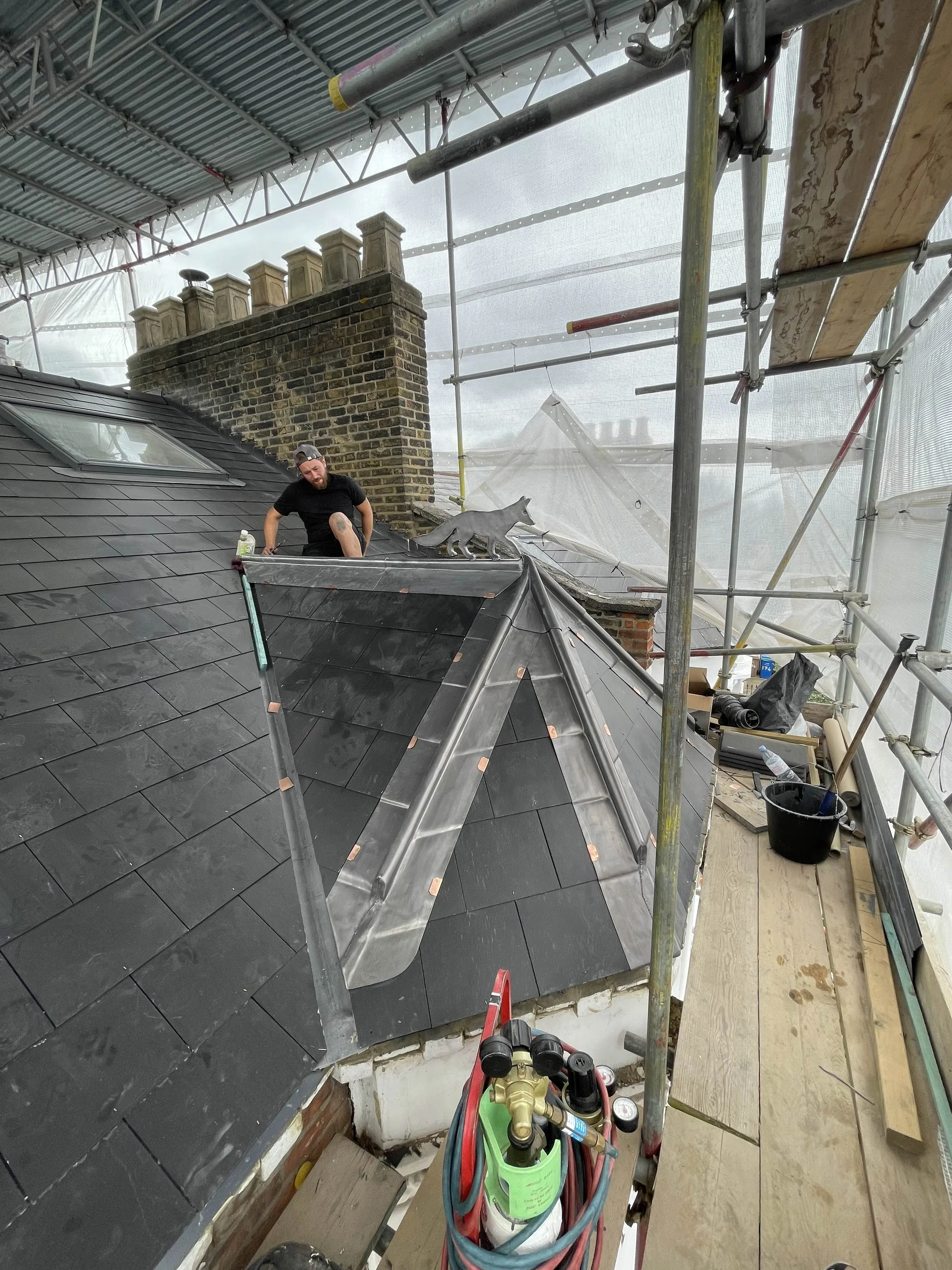 A worker on a rooftop construction site installing or repairing black shingles and metal flashing, surrounded by scaffolding and construction tools.
