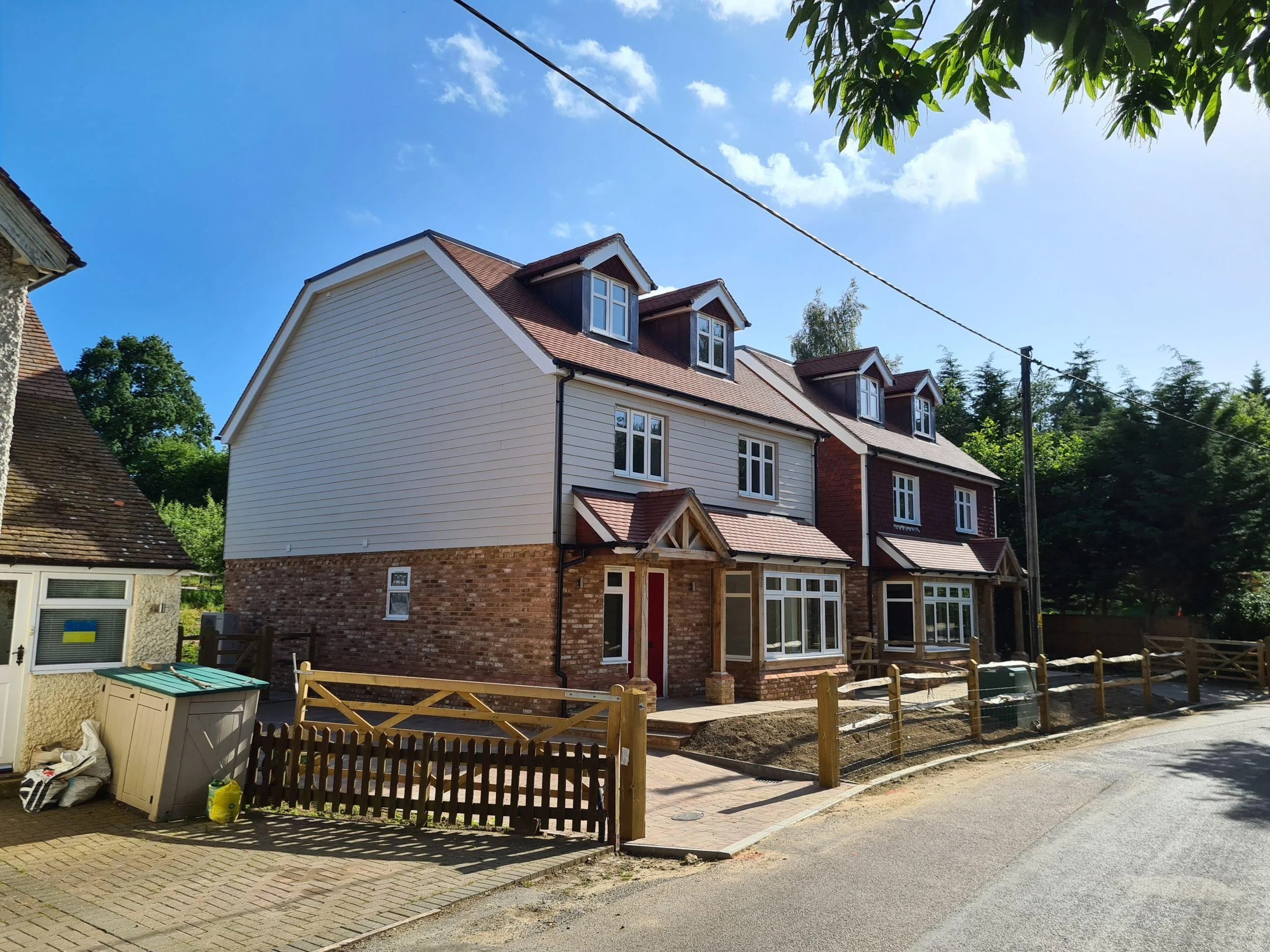 Newly constructed three-story house with brick and white siding exterior, multiple windows, and a small porch, surrounded by a wooden fence, set on a street under a partly cloudy sky.