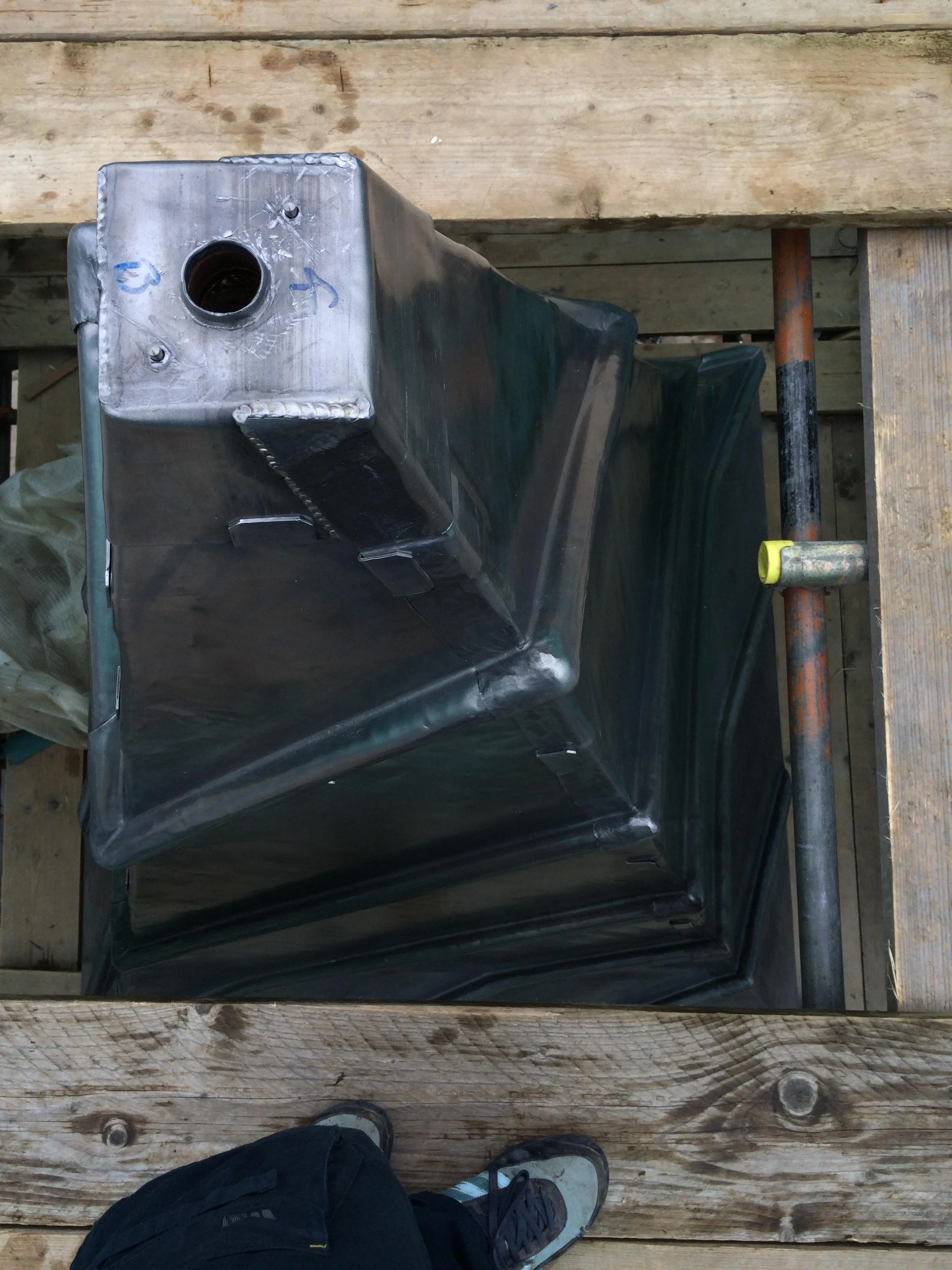 Metal tank with a large opening at the top, placed on a wooden platform with a wooden railing and support beams around.