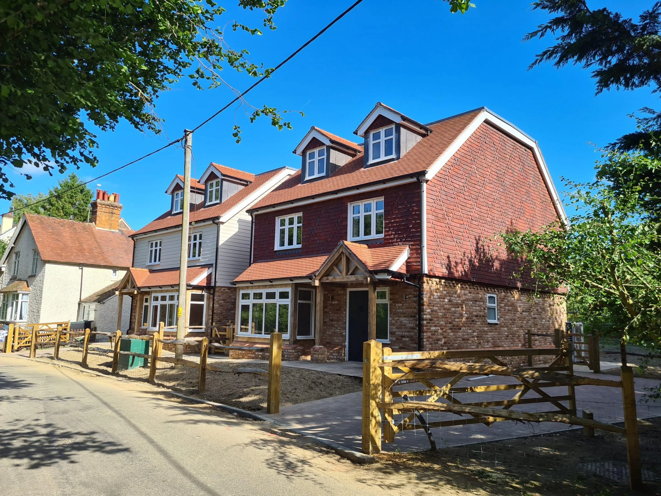 Newly built, multi-story house with a brick lower level, white and red siding upper level, and multiple roof dormer windows, situated along a street with a wooden fence and trees.