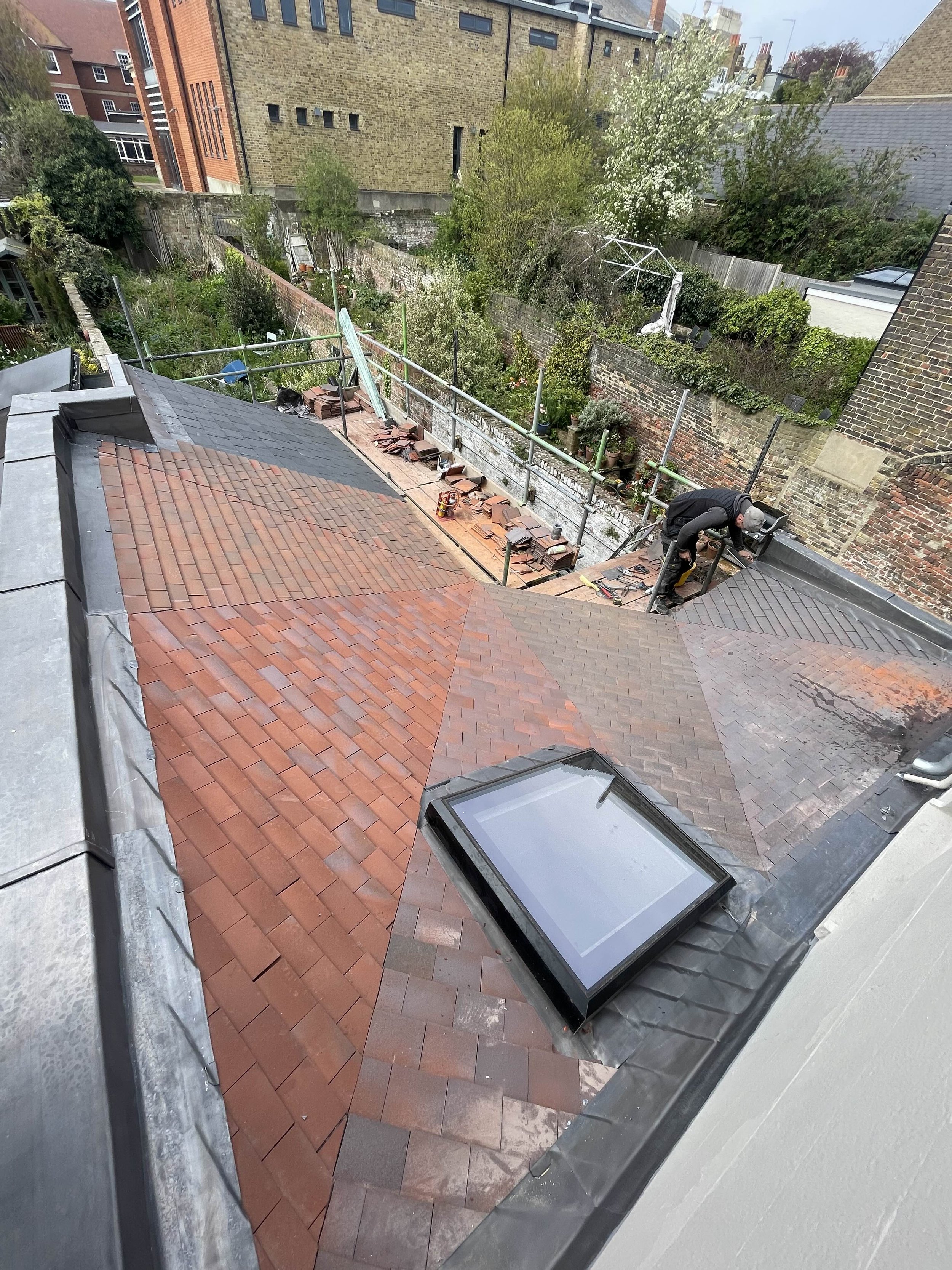 A rooftop with new and partially completed roof tiles in multiple colors, including reddish-brown and darker shades. A worker is installing tiles near a skylight window, and there are construction materials and tools on the roof.