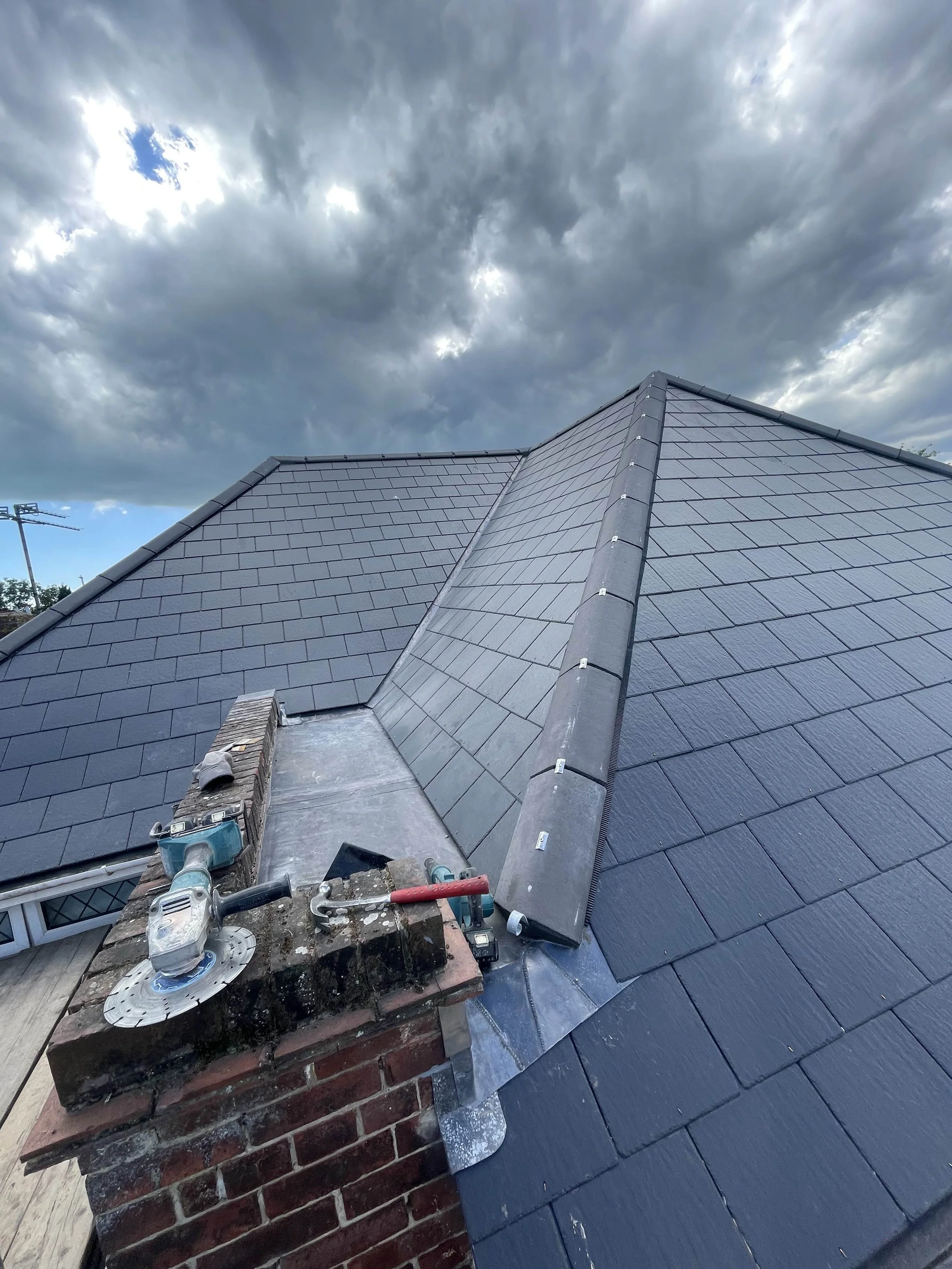 Black slate roof nearing completion under stormy cloudy sky, with tools and materials on chimney brick in the foreground.