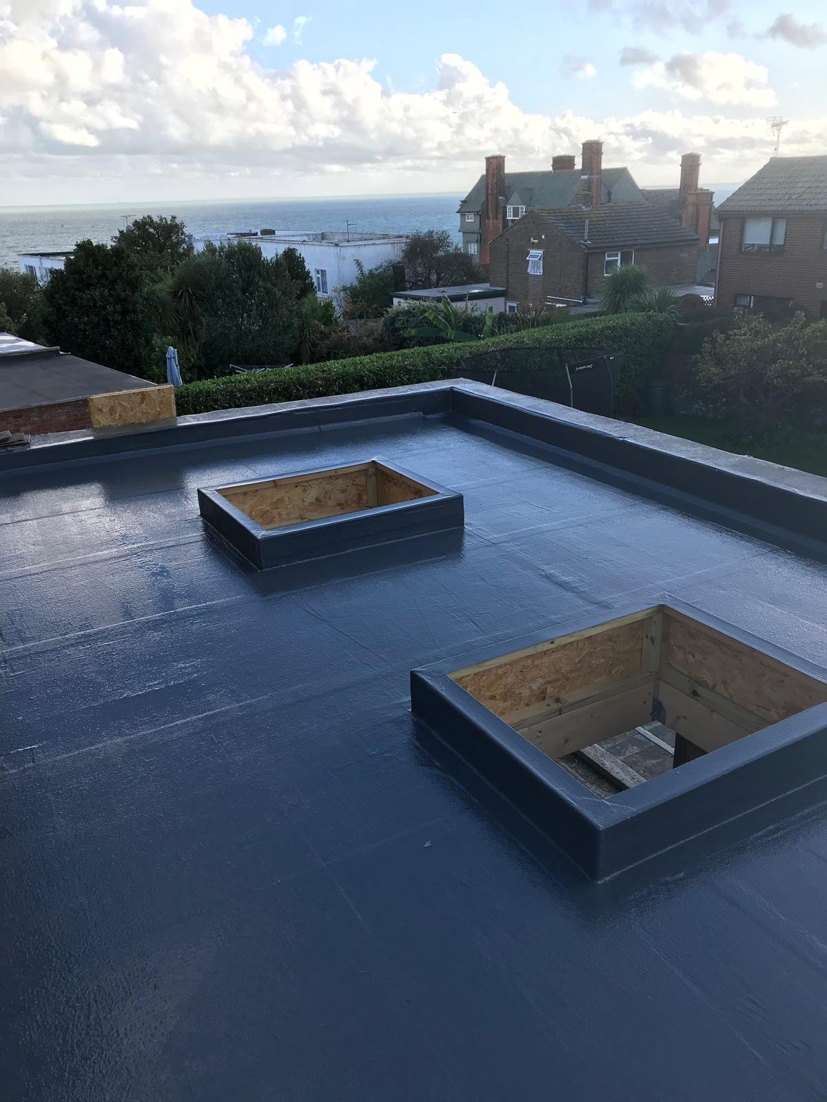 View of a flat roof under construction with two rectangular openings, overlooking houses, trees, and the ocean in the distance under a partly cloudy sky.