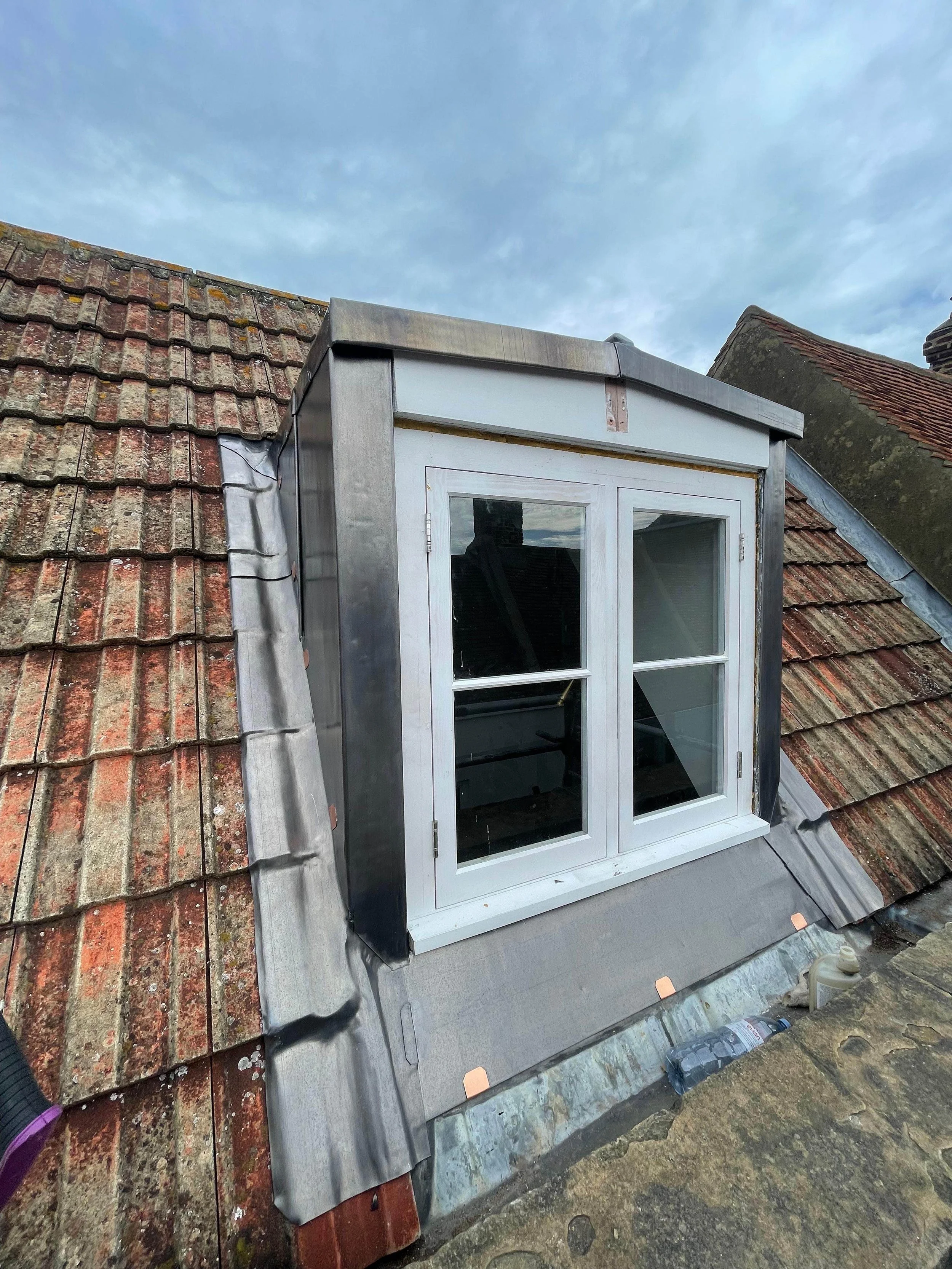 Exterior view of a roof installation with a white dormer window, metal flashing, and old clay tiles, under a cloudy sky.