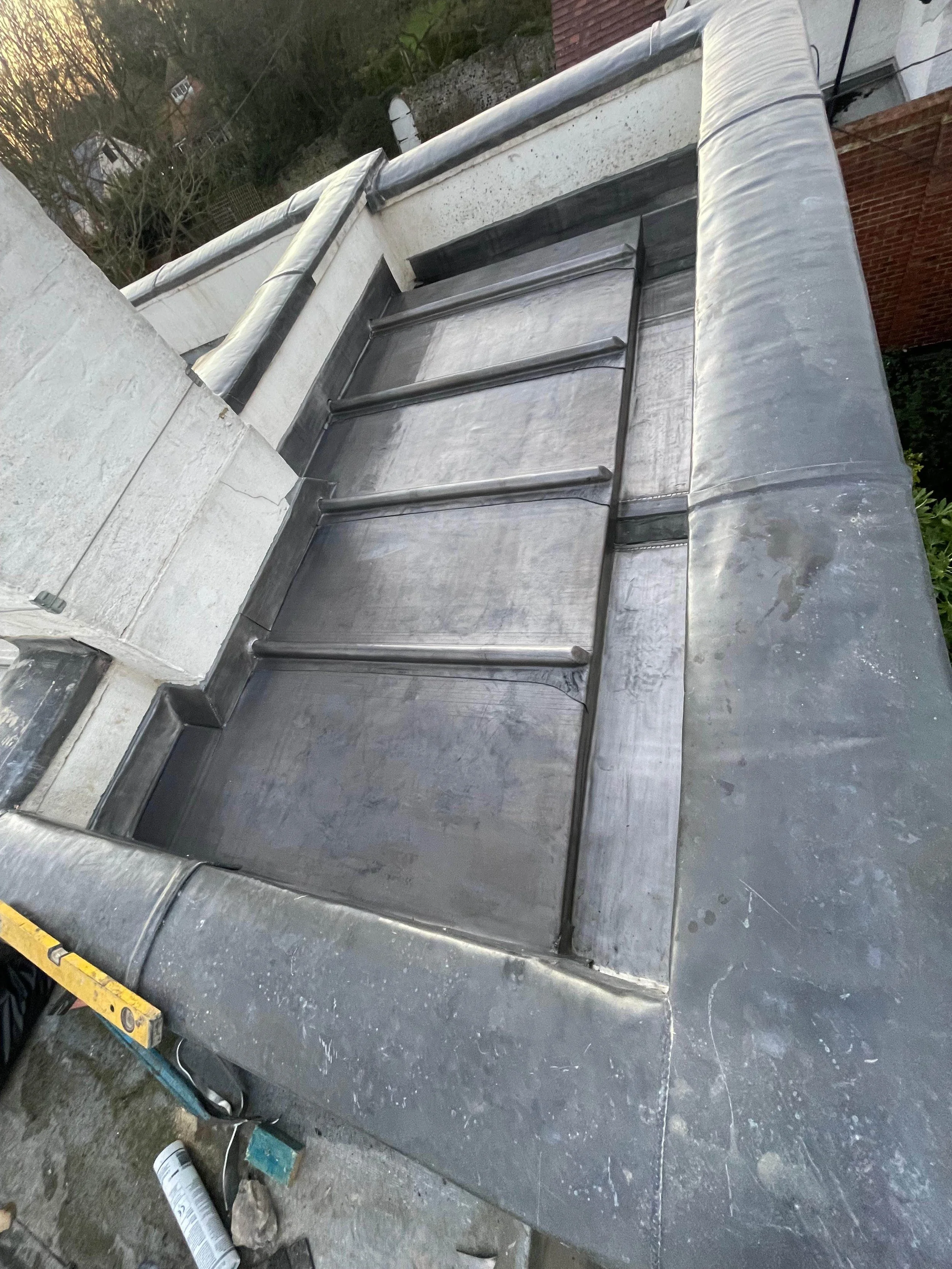 View of a flat rooftop with metal framing and flashing, possibly during construction or repair, with trees and neighboring houses visible in the background.