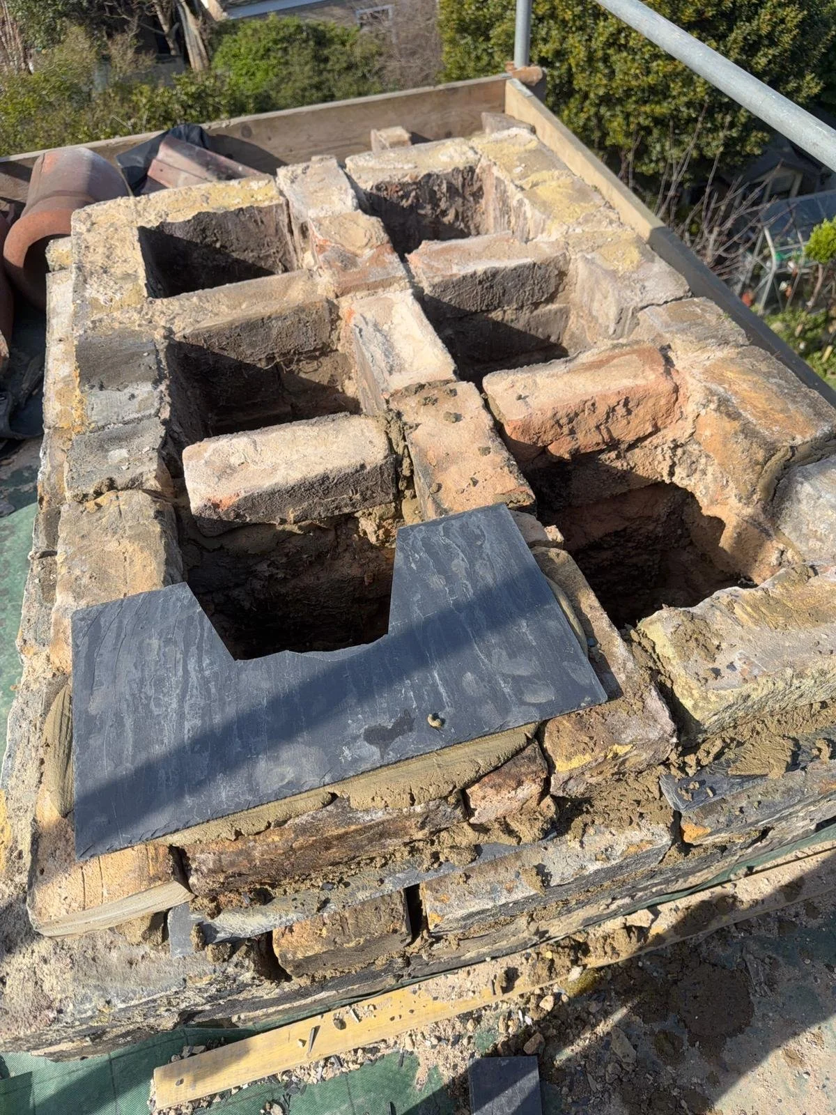 Brick chimney with an opening at the top, with some bricks removed, on a scaffolding or platform outdoors.