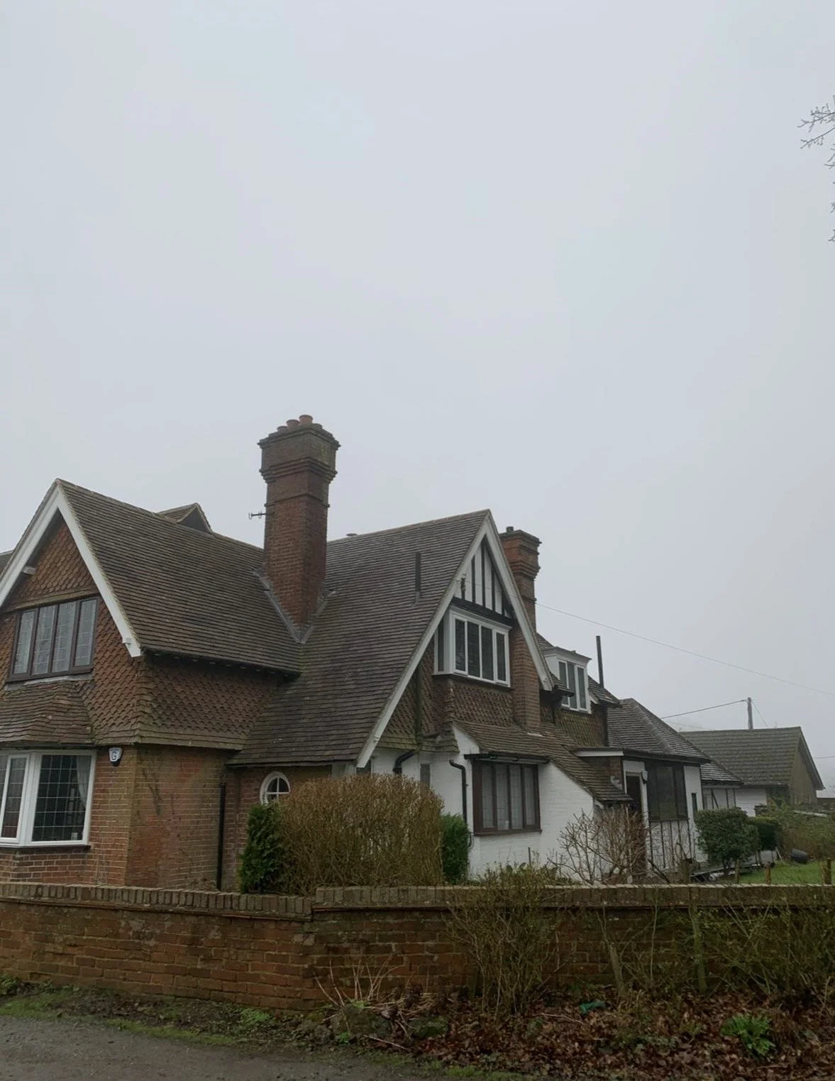 A large, traditional brick and white house with a steep roof, multiple chimneys, and various windows, surrounded by a brick wall and some bushes, on a cloudy day.