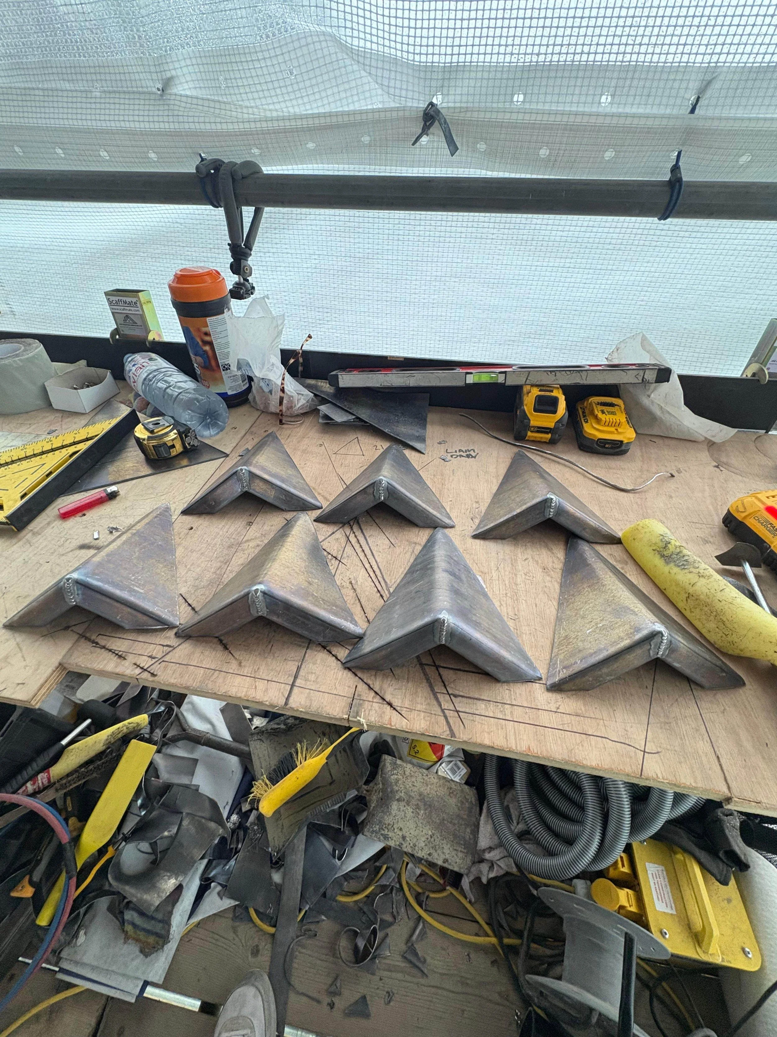 Workshop table with metal pyramid-shaped constructs, tools, measuring tape, level, rubber mallet, and various construction supplies, with window in background.