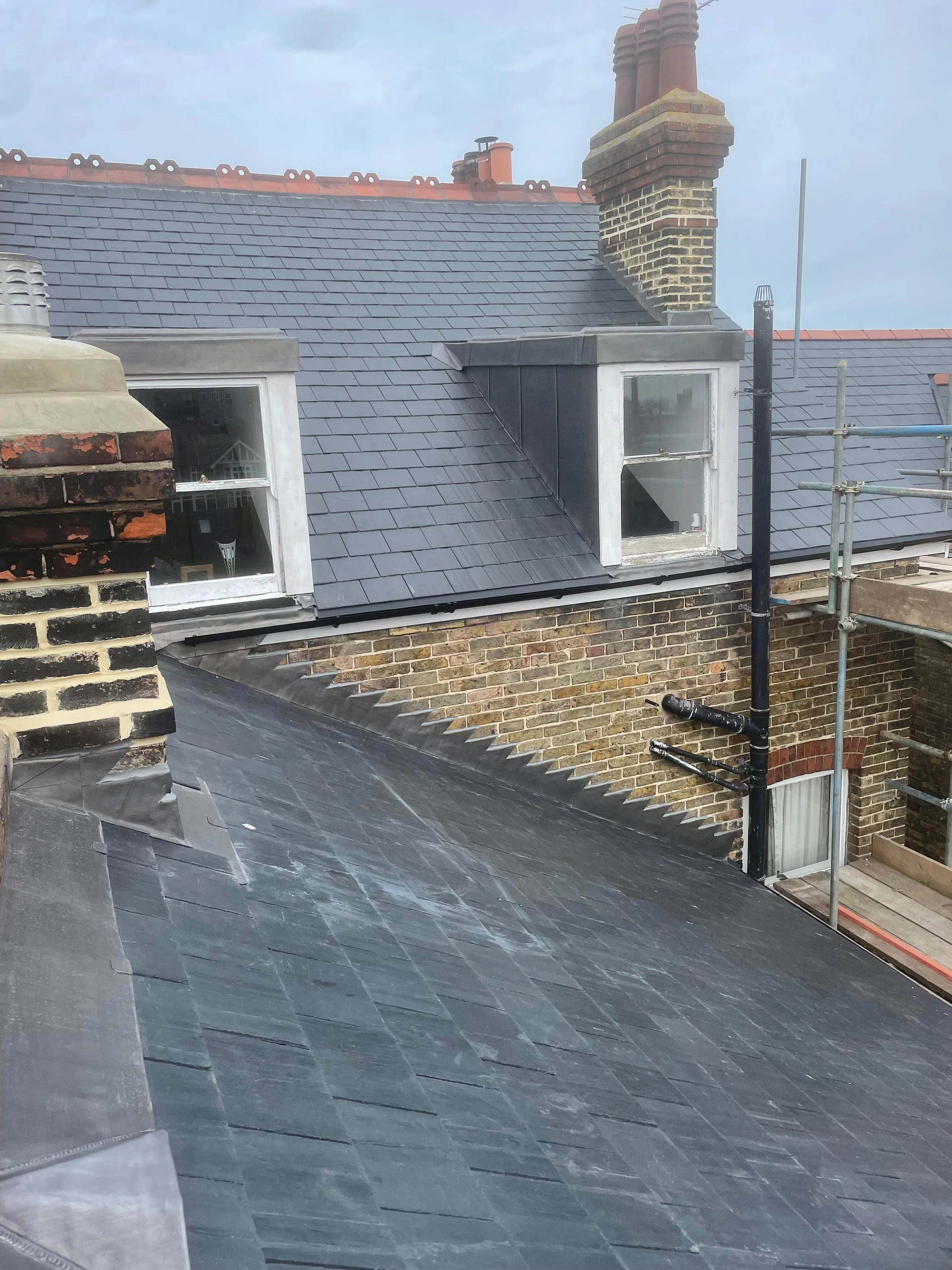 View of rooftops with slate shingles, brick chimneys, and dormer windows on a cloudy day.