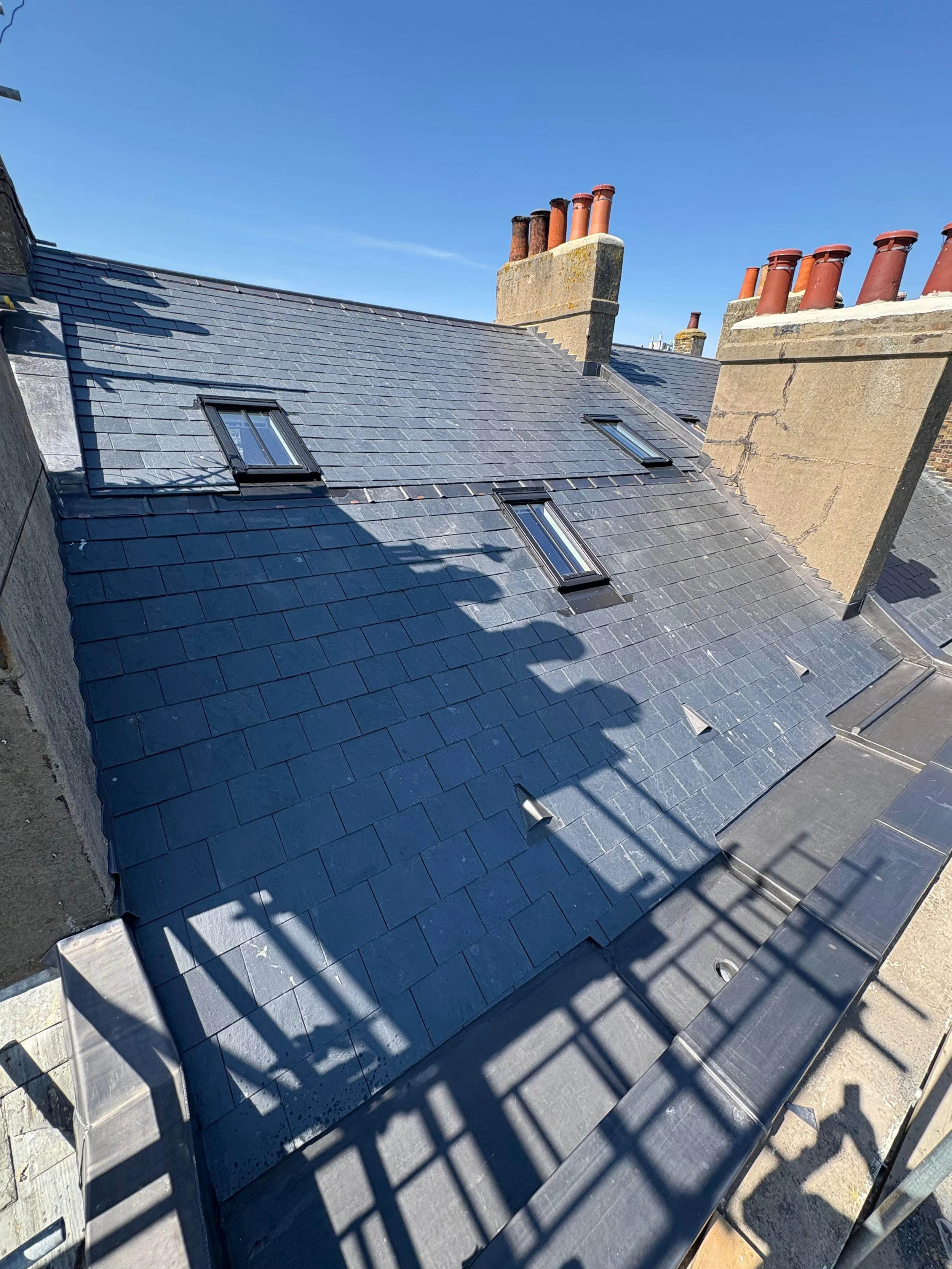 View of a rooftop with dark slate shingles, three skylights, and multiple chimney stacks with red clay pots under a clear blue sky.