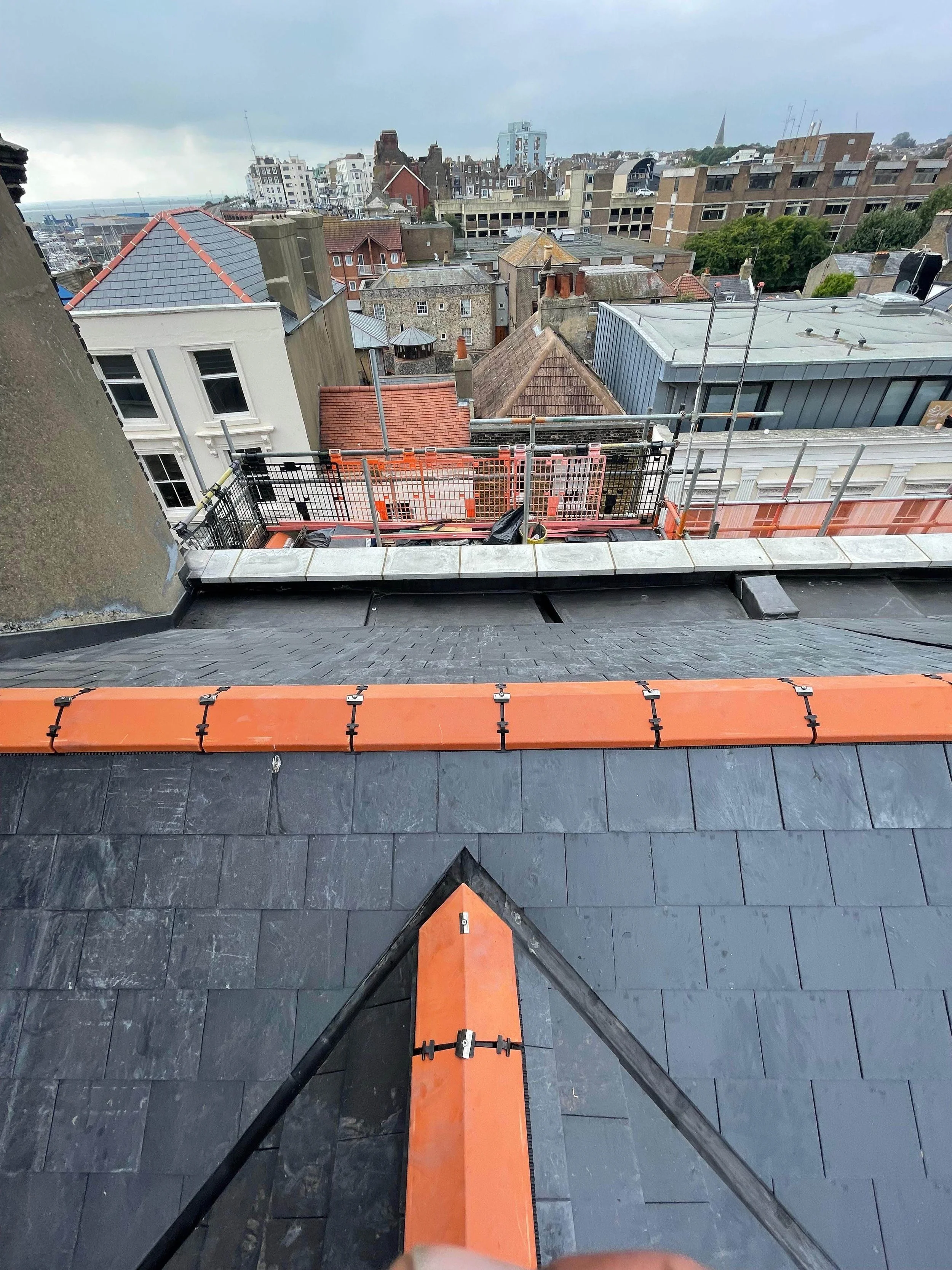 View from a rooftop construction site showing chemical weathered slate roof tiles, safety railings, and a cityscape with various buildings and cloudy sky in the background.