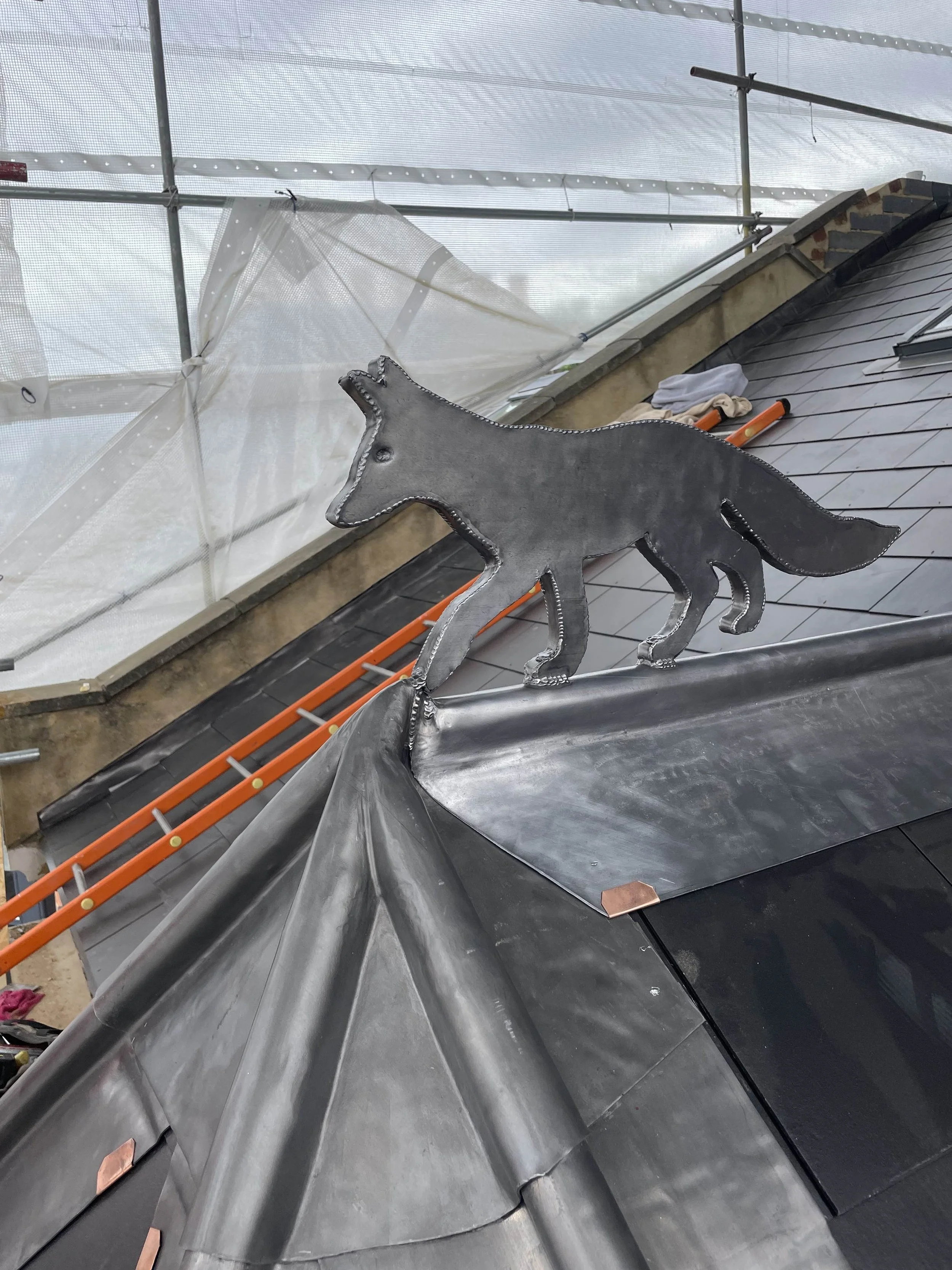 Metal wolf sculpture perched on a rooftop ridge during construction, with scaffolding and roofing tools visible in the background.