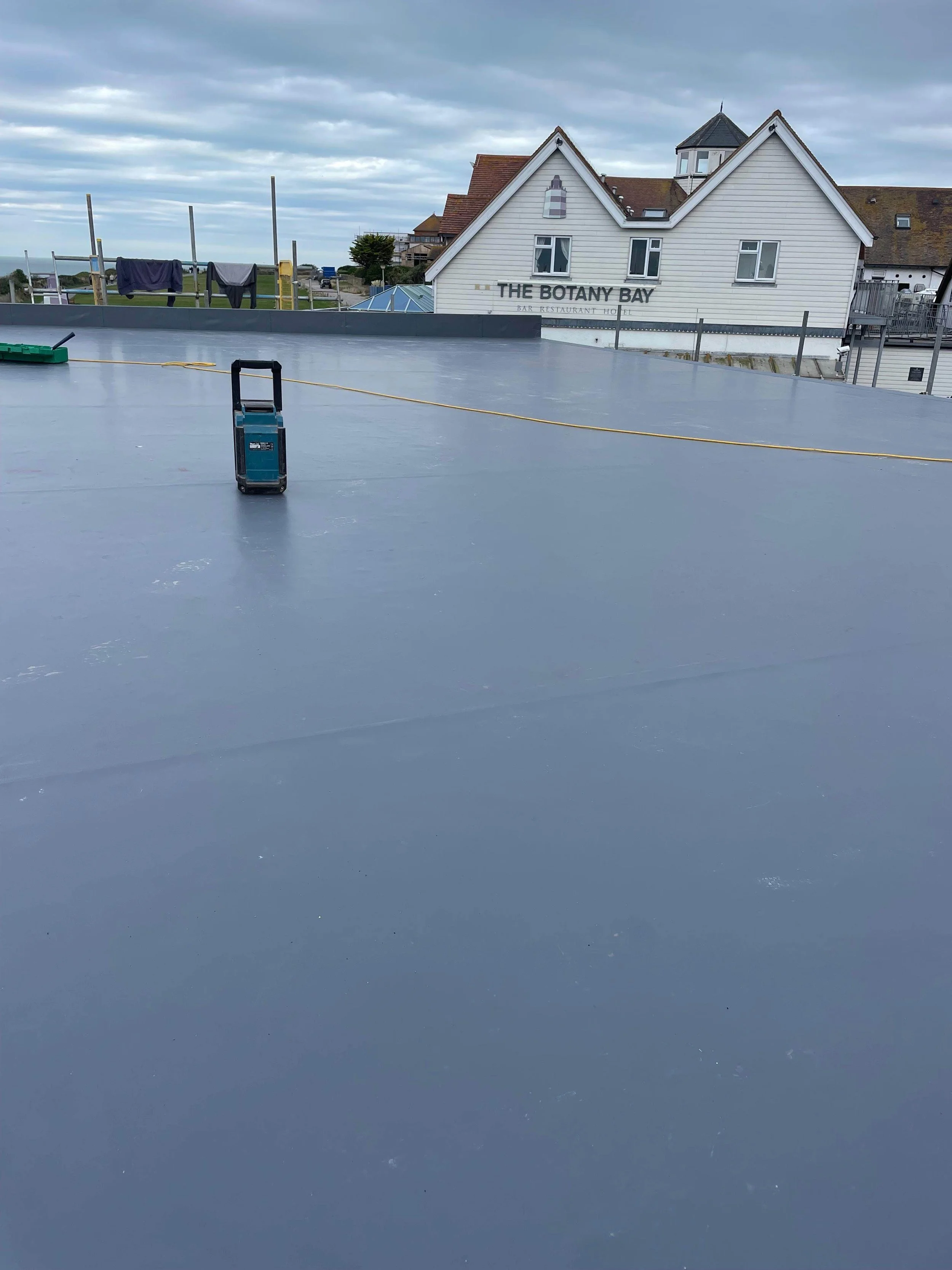 Empty outdoor rooftop with gray surface, some equipment, and a white building with a sign that reads 'The Botany Bay' in the background under a cloudy sky.