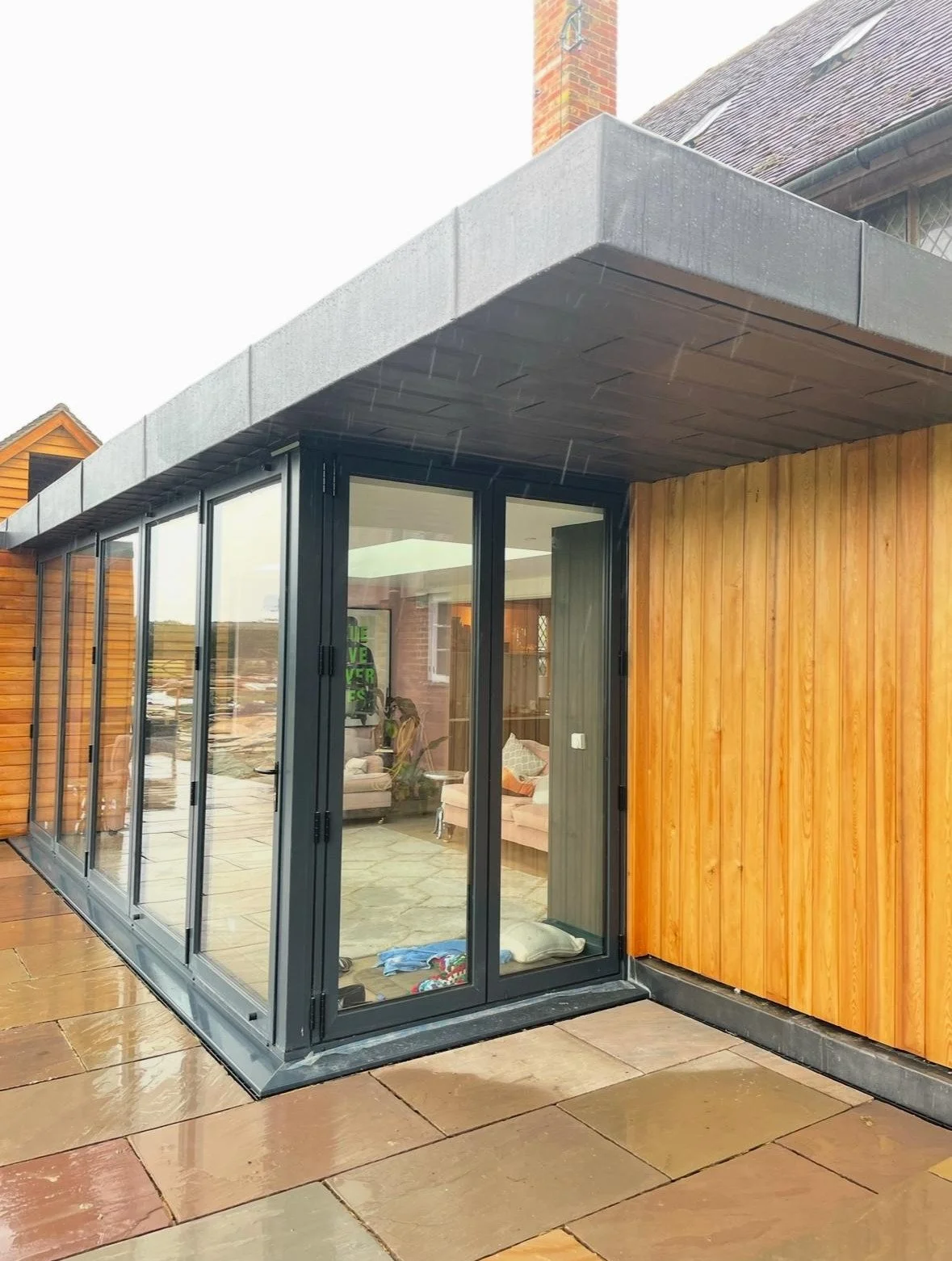 Exterior view of a modern building with wooden siding, black framed glass doors, and a flat gray roof overhang on a wet tiled patio.