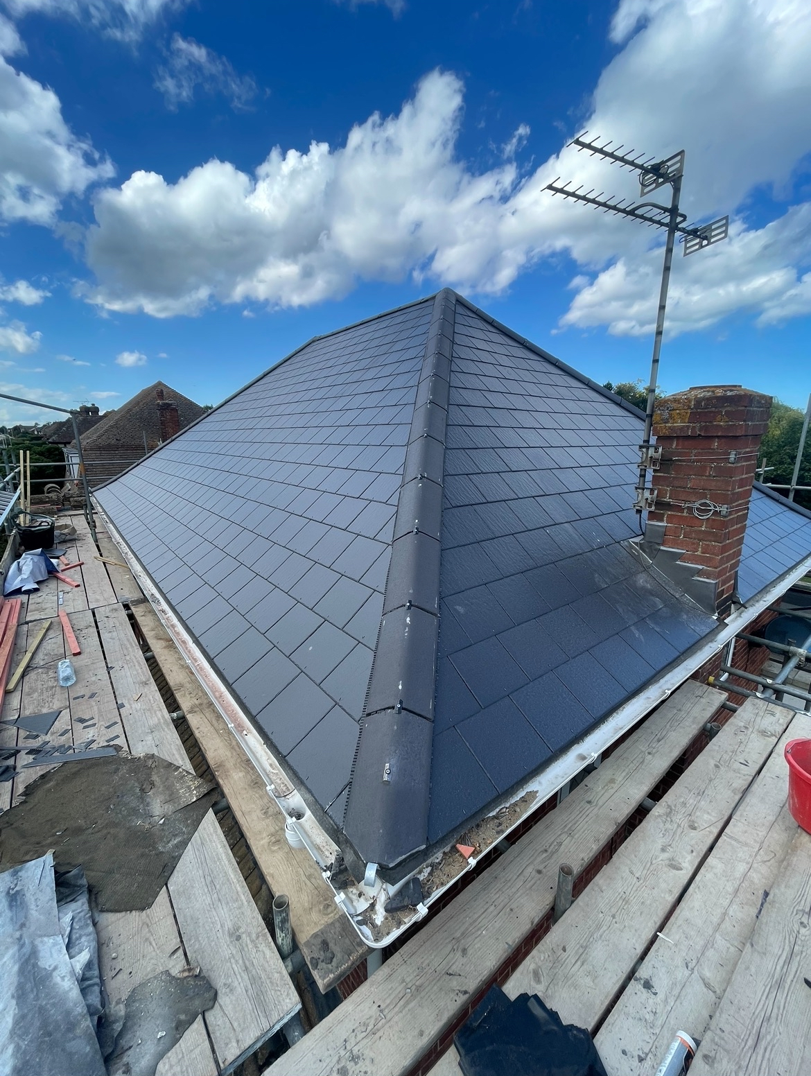 Newly installed dark grey tiled roof on a building under construction with scaffolding, a chimney, and a TV antenna against a blue sky with white clouds.