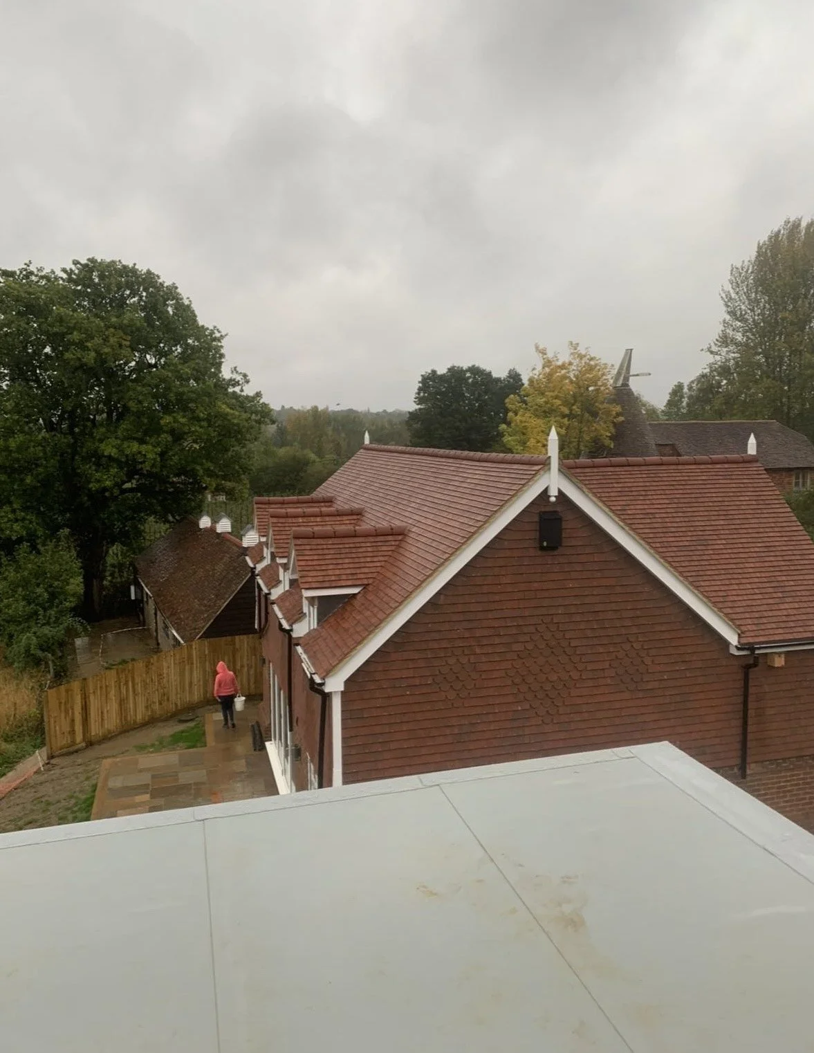 Overcast day view of residential rooftops, trees, and a person in a red hoodie walking near a fence.