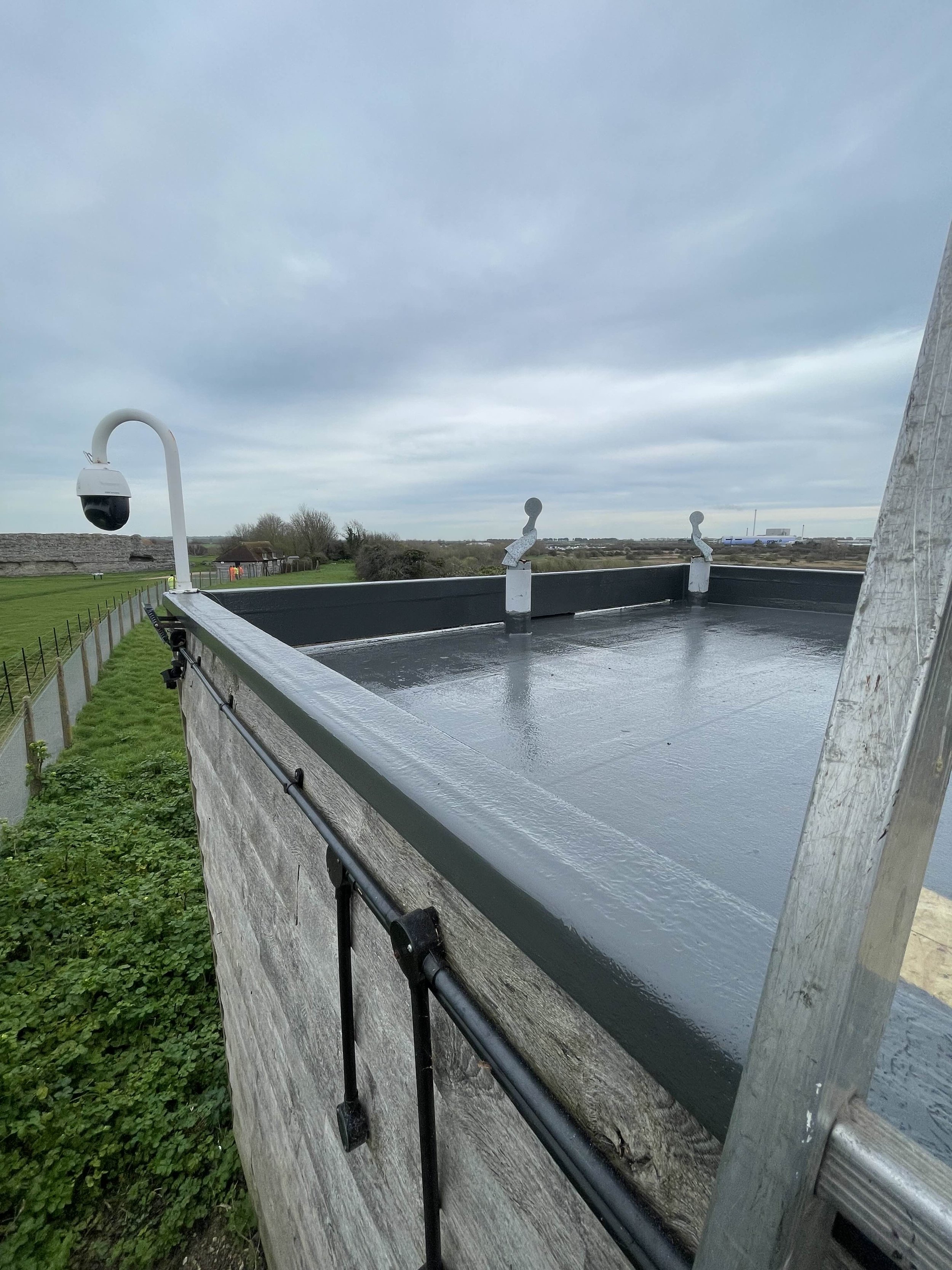 Rooftop with surveillance camera and two weather vanes, with cloudy sky and landscape in the background.