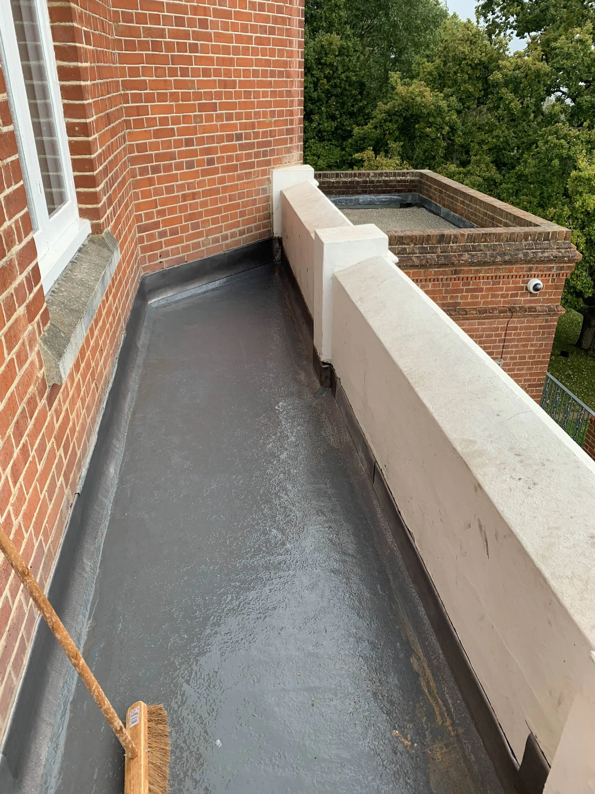 A balcony with a black waterproof floor, a brick wall with a window, a white wall with a barrier, and a view of trees in the background.