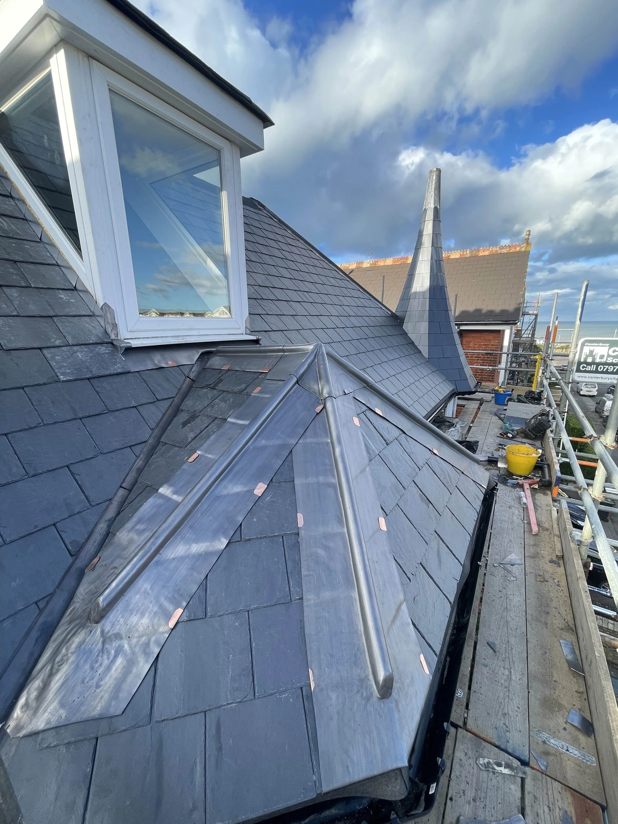 View of a roof with slate tiles and metal flashing, part of a construction or renovation project on a house, with scaffolding and construction tools visible.