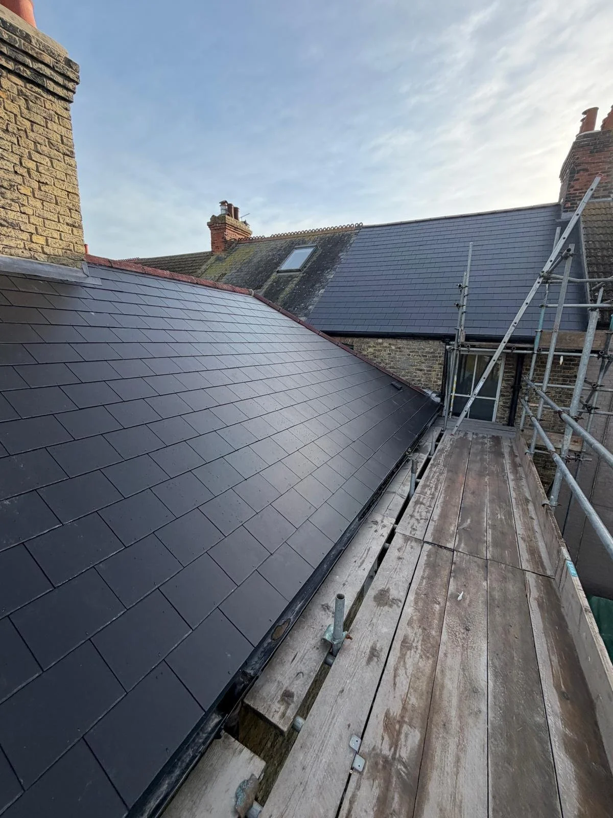 Photo of a rooftop with new black tiles, adjacent to scaffolding and a wooden platform for construction work.