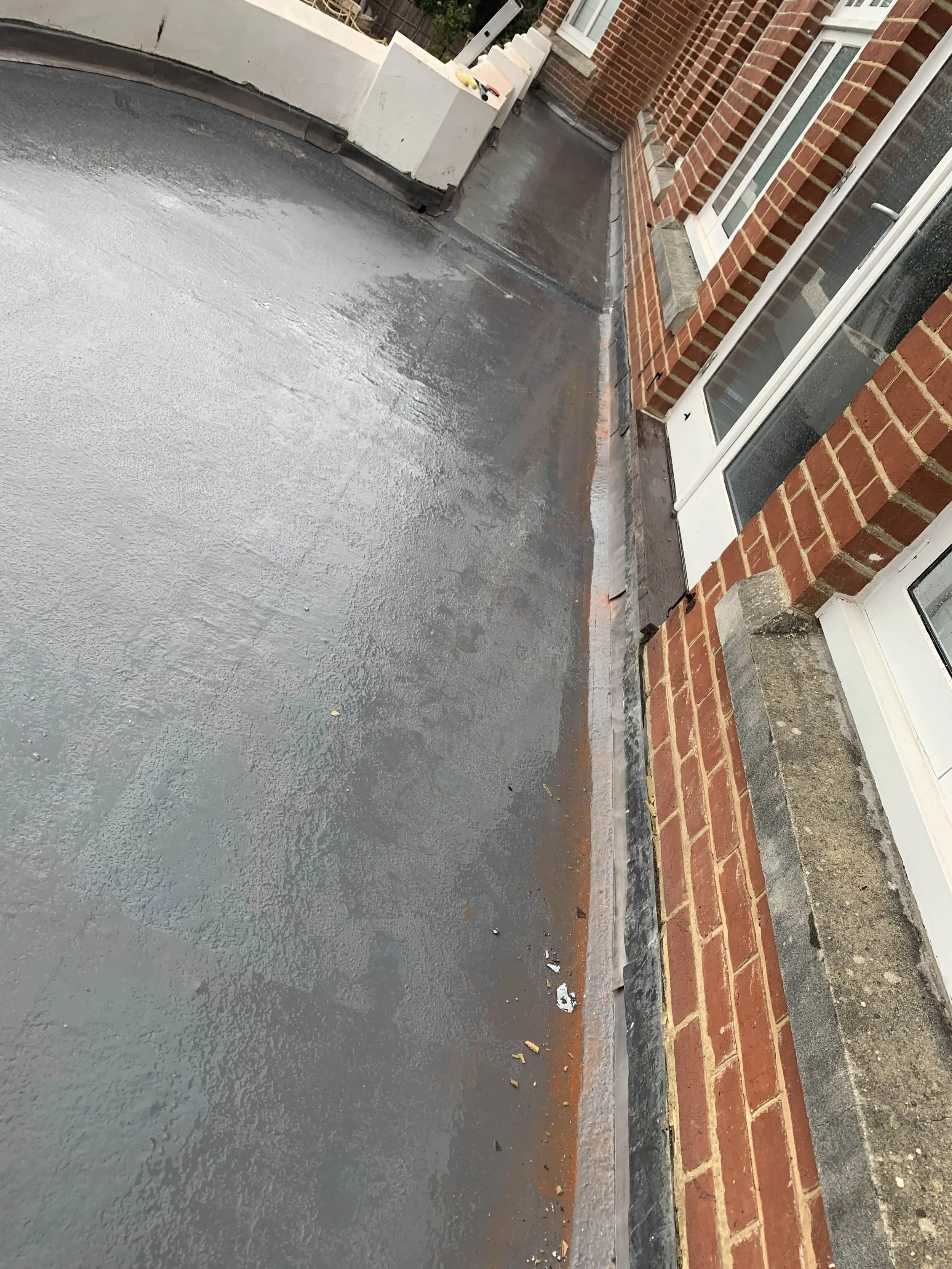 View of a flat roof with black waterproof coating next to a red brick wall with white window frames.